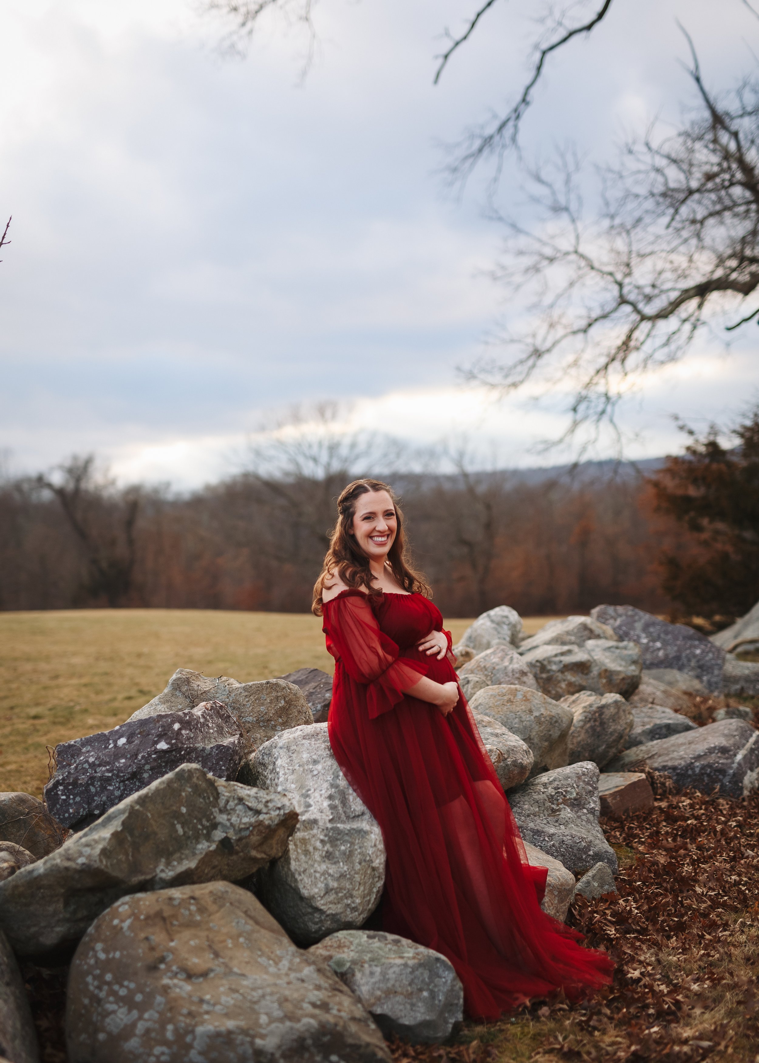  Mother-to-be in an elegant red maternity gown leaning against a rock wall in a New Jersey field 