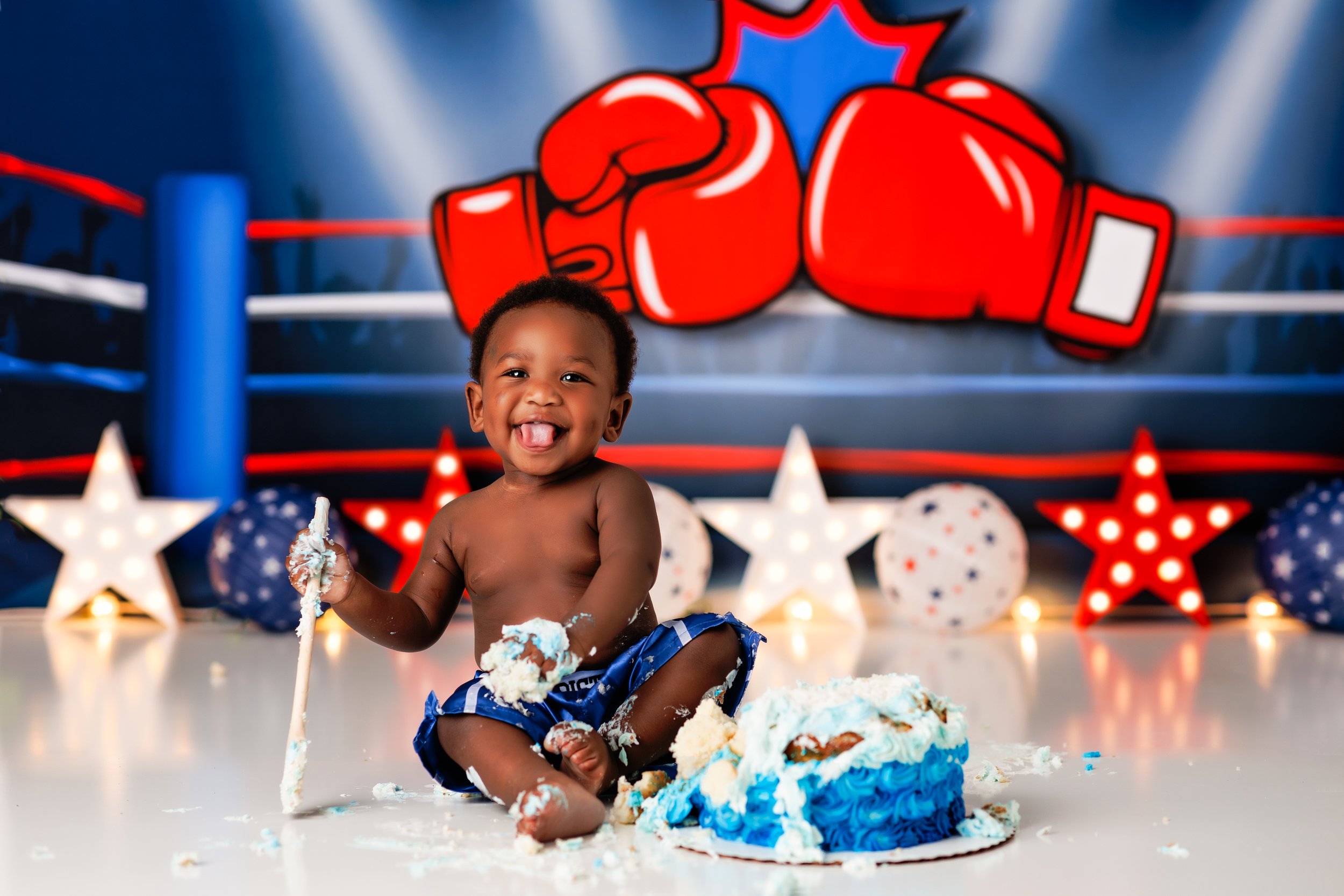  Black baby holding a wooden spoon as he prepares to eat his birthday cake, with a boxing glove banner behind him 