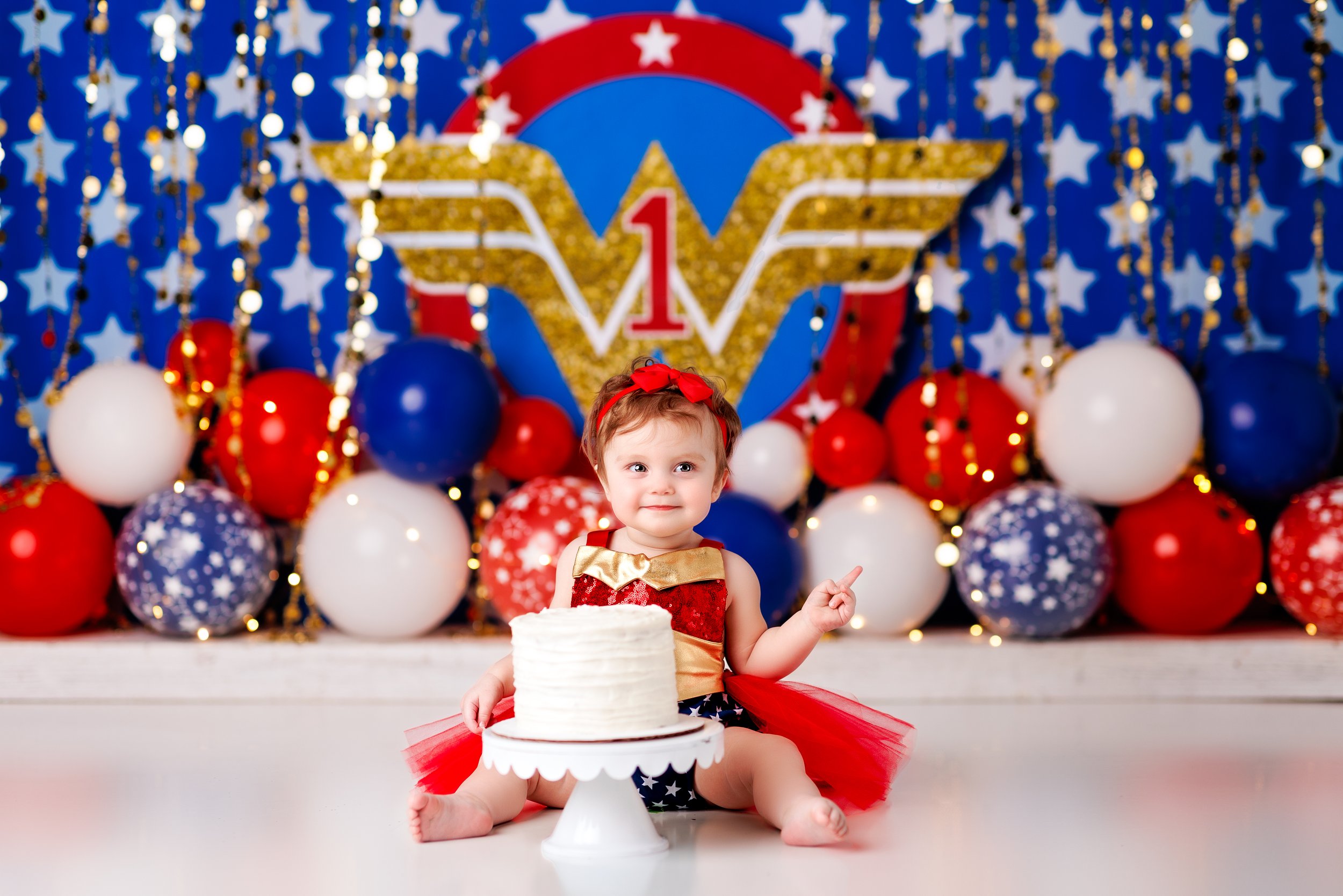 Baby in red and blue dress with red, white, and blue balloons and the Wonder Woman logo behind her  
