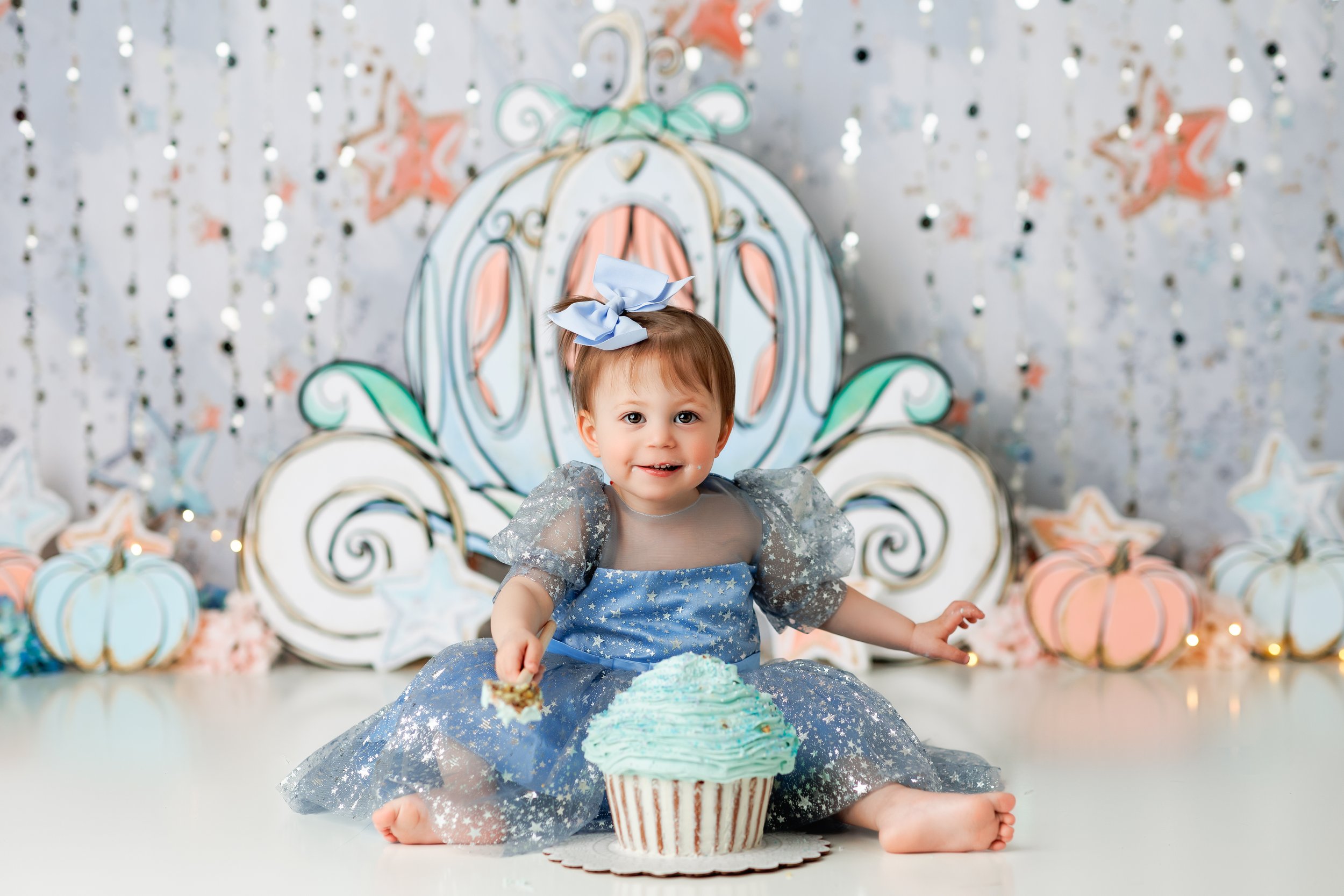  Baby girl in blue dress eating her cake with a carriage in the background during her Cinderella themed birthday photoshoot 
