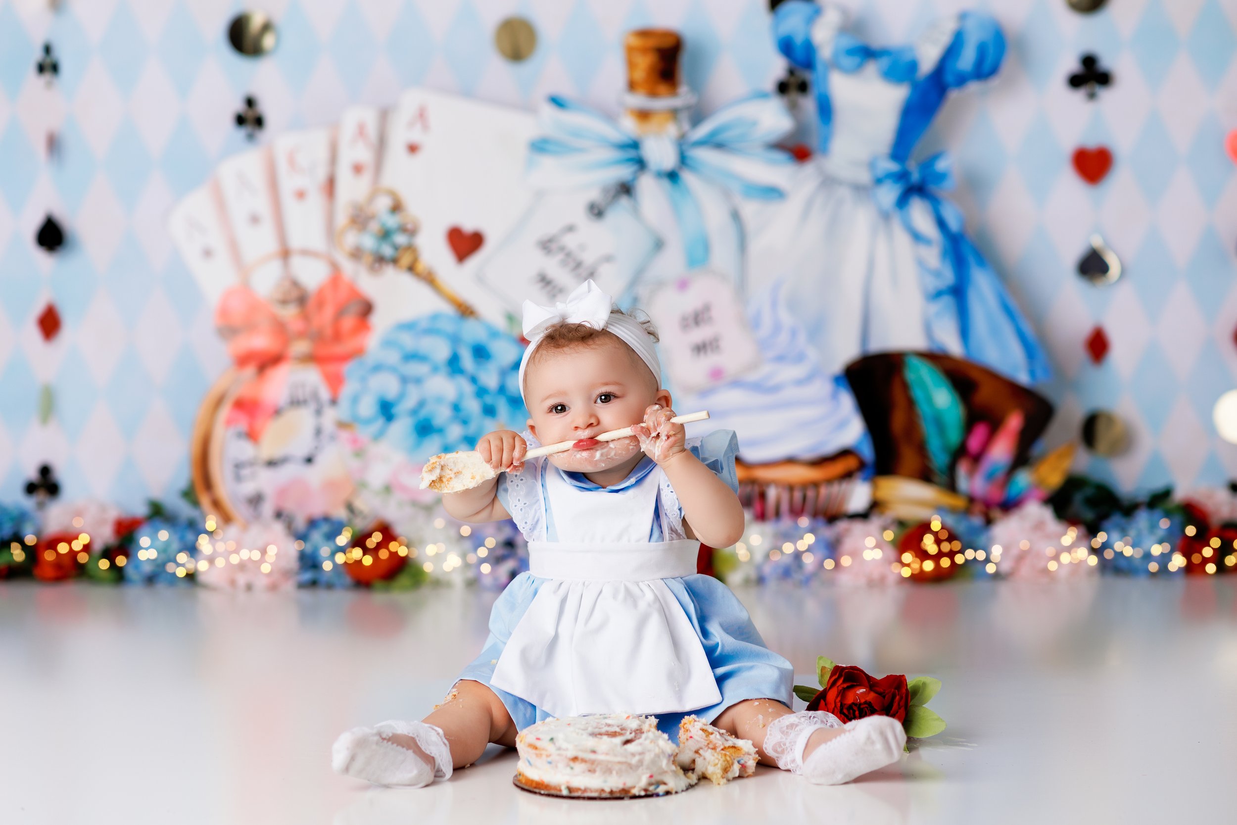  Baby girl in blue dress chews on a wooden spoon that she uses to eat her cake against an Alice in Wonderland backdrop 