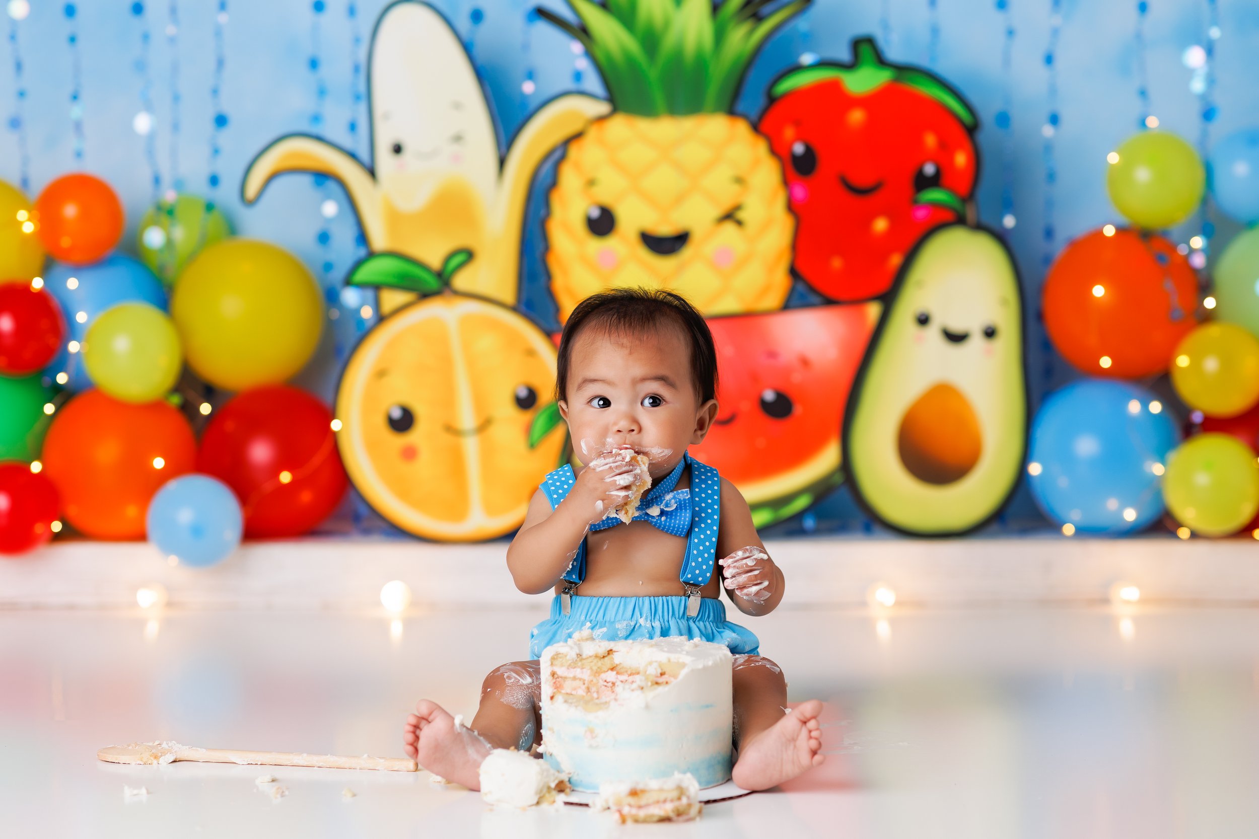  Baby boy stuffs cake into his mouth during his first birthday photoshoot against a backdrop of tropical Kawaii fruits 