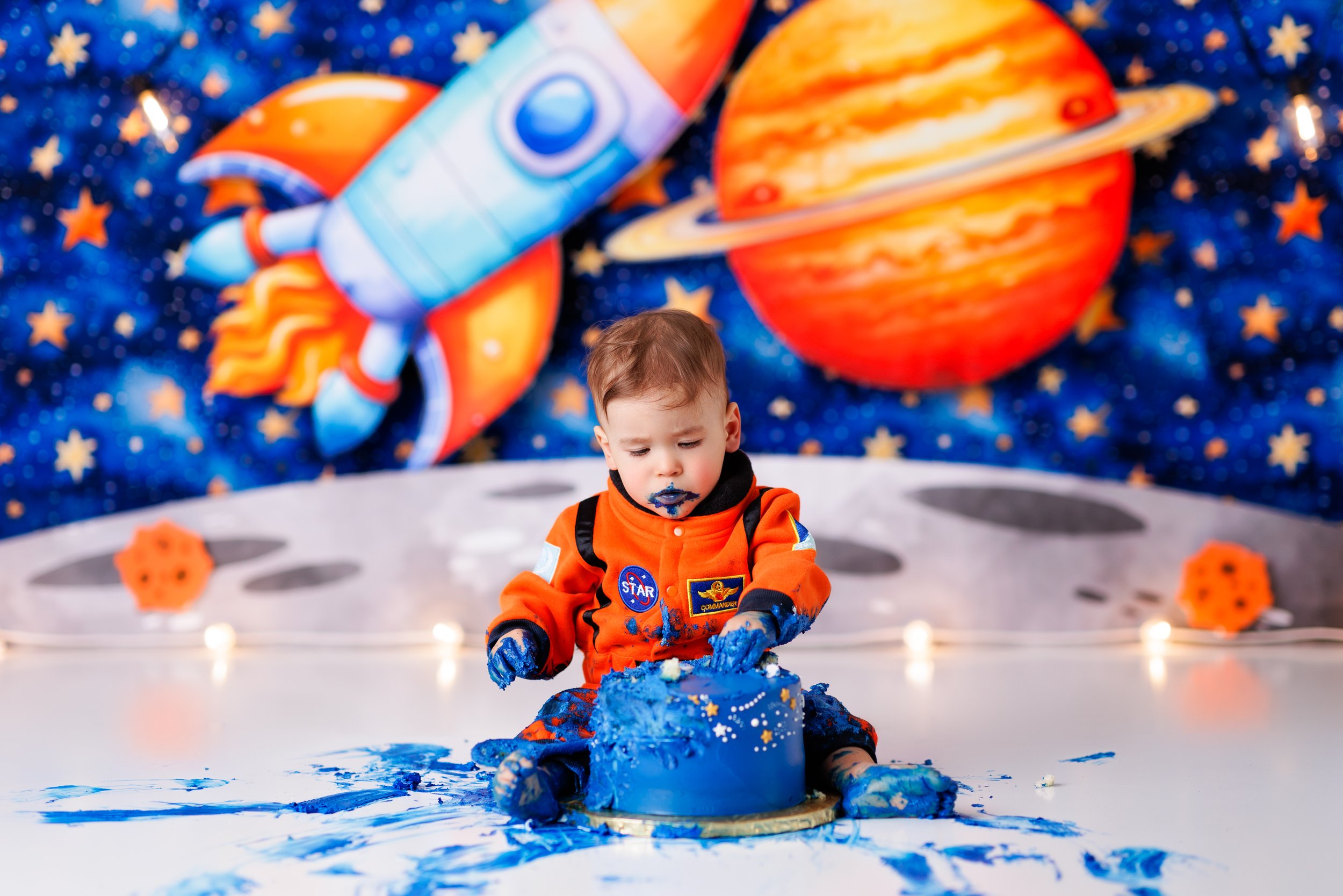  Toddler boy in astronaut suit dives into his blue cake with a moon and planets backdrop 