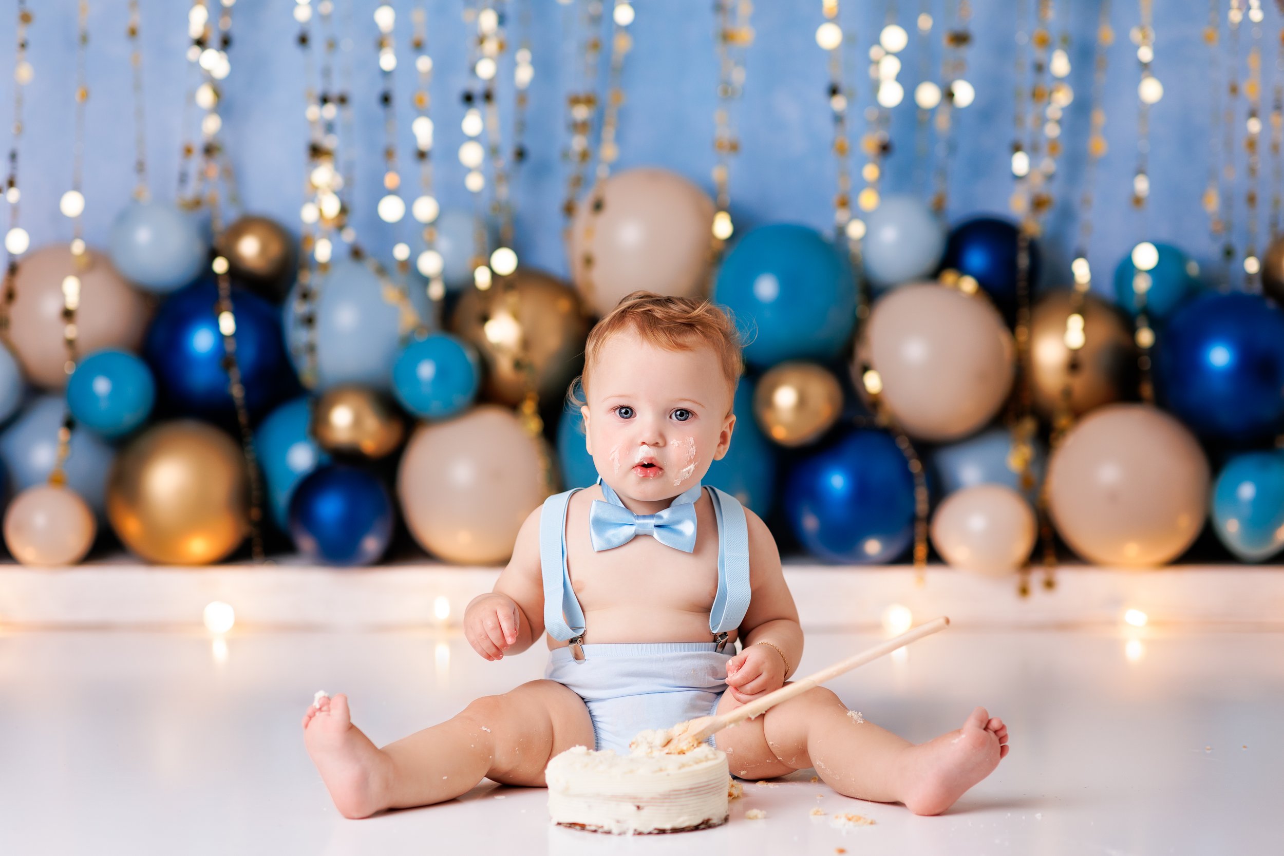  Baby boy in blue sitting by his birthday cake with blue and gold balloons behind him 