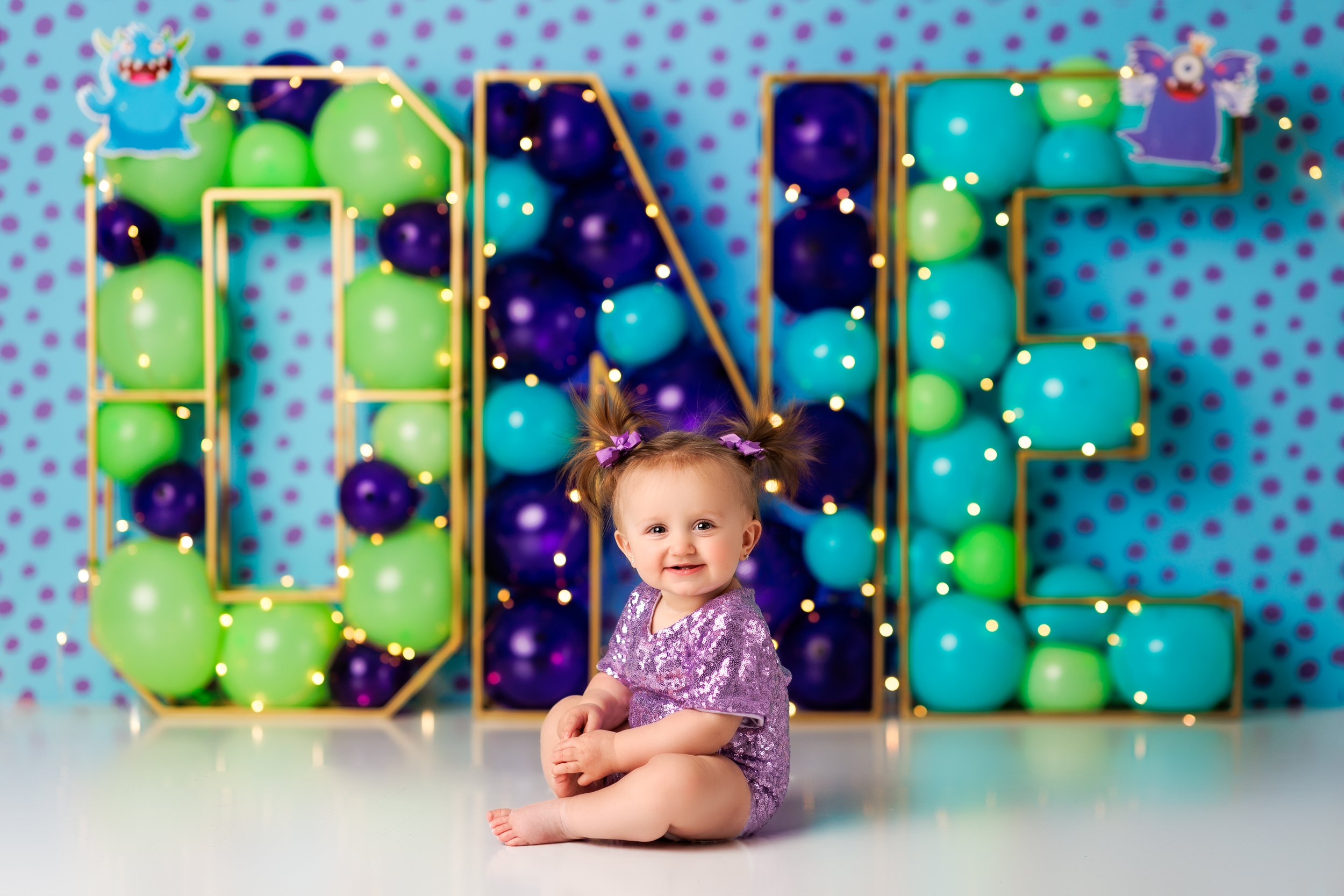  Cute baby girl in purple seated in front of balloon frame spelling out the word “one” 