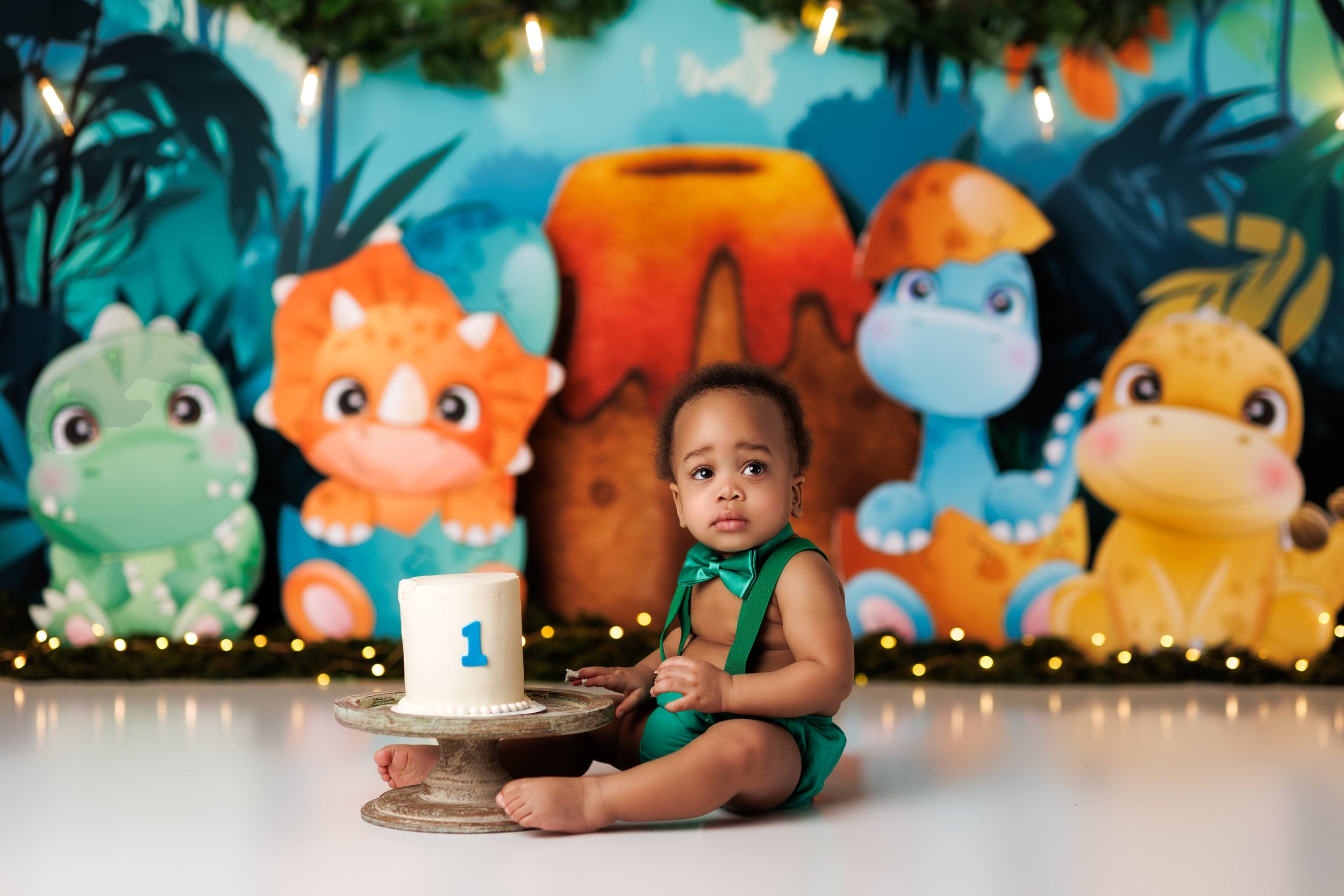  Cute black baby seated in front of a dinosaur friends backdrop for his birthday cake smash 