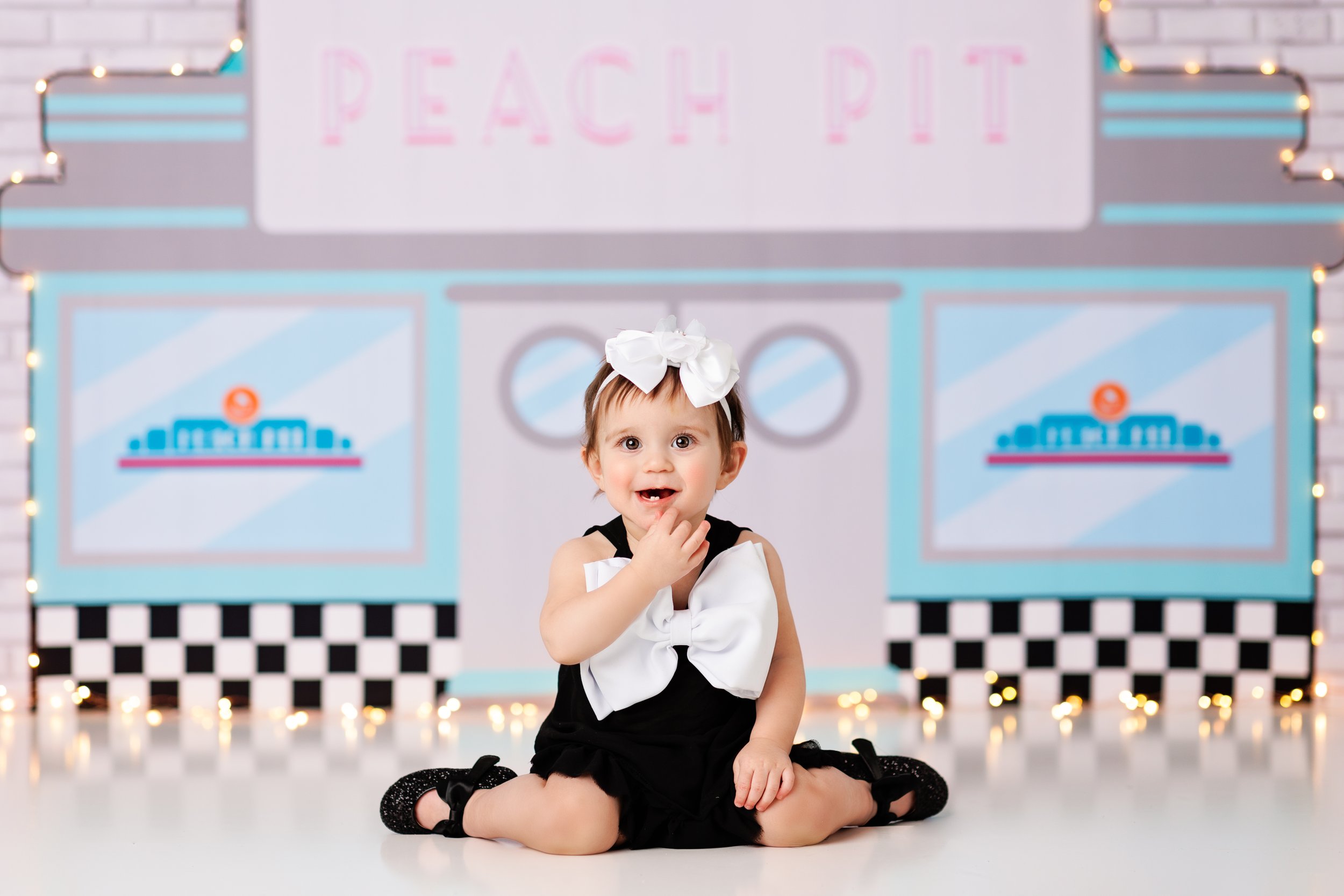  Baby girl in black dress with big white bow seated in front of a Peach Pit sign during her 90210-themed cake smash 