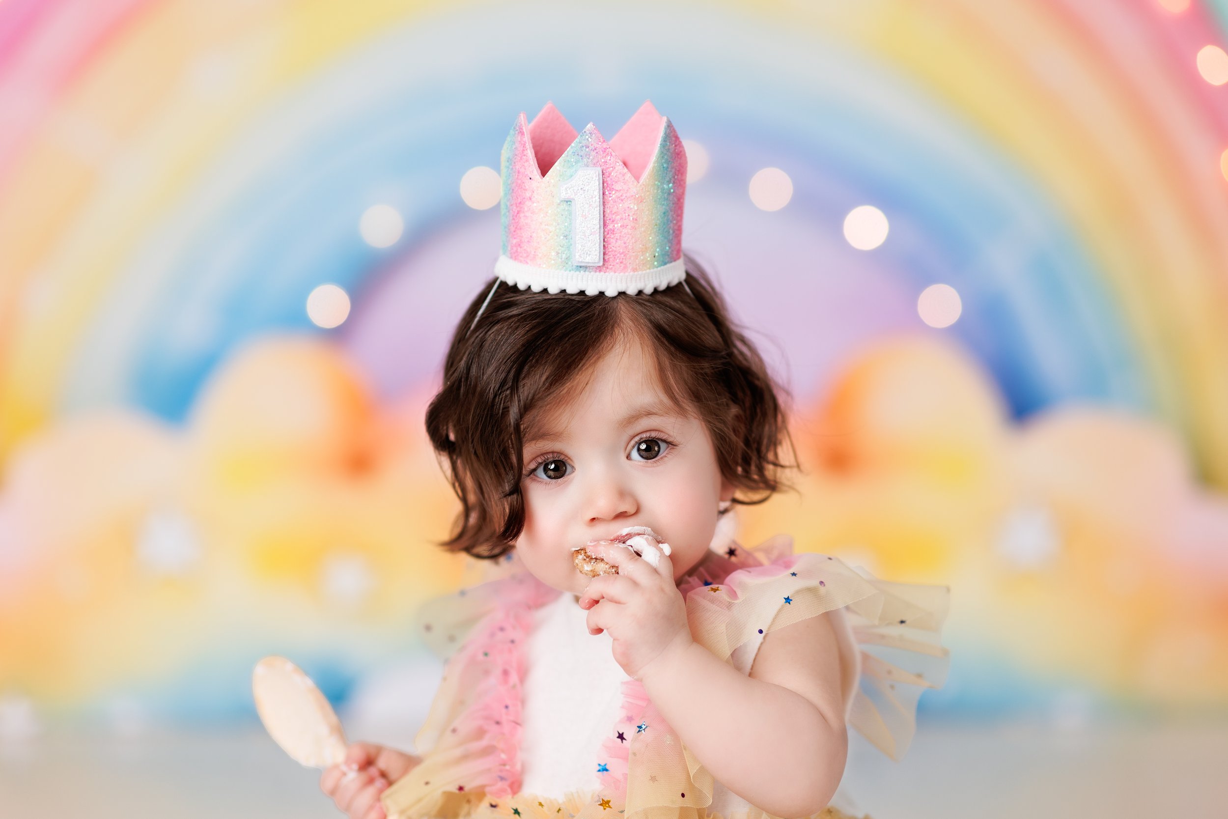  Toddler girl wearing a crown and licking icing off her fist during a rainbow themed cake smash 