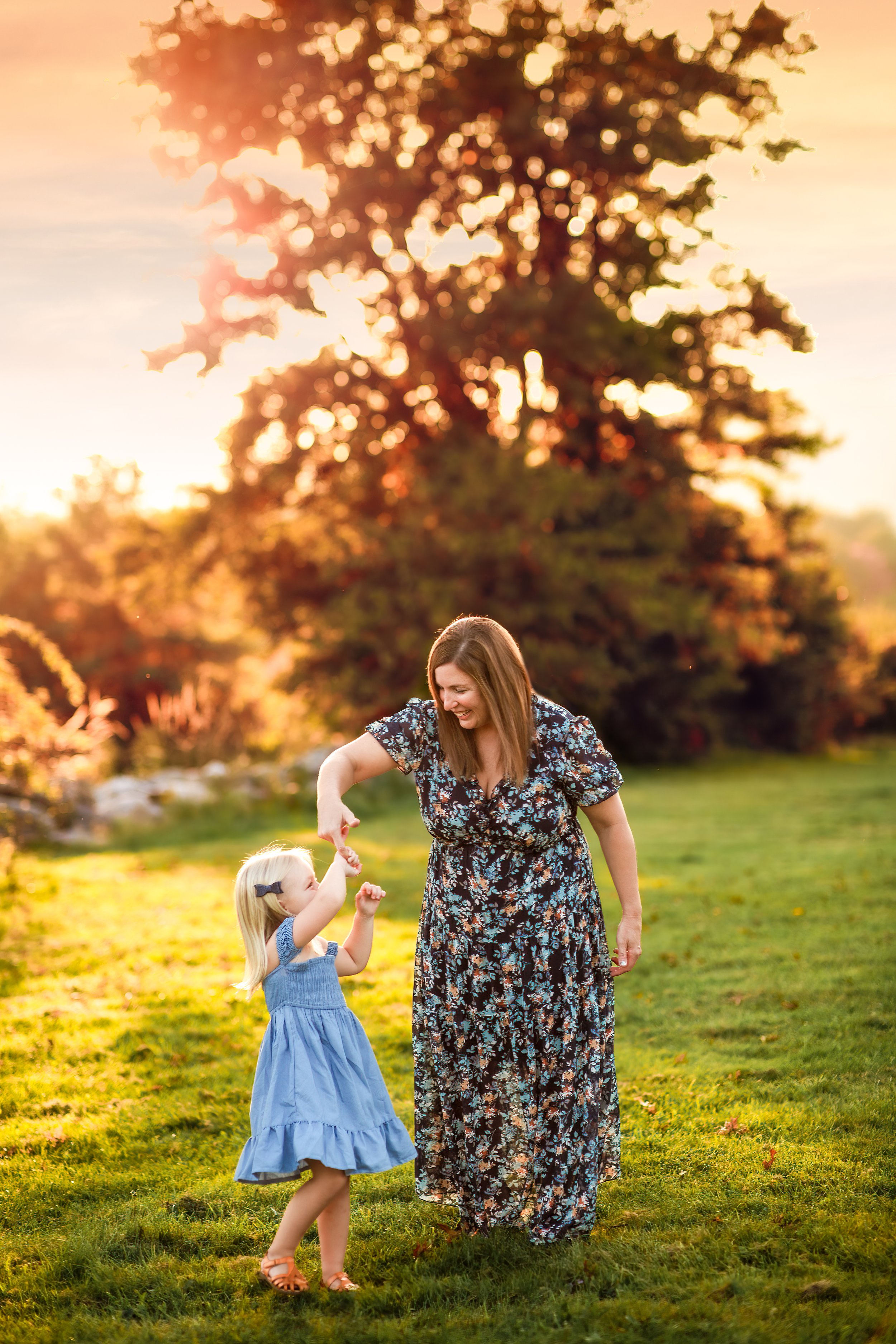  Mother in floral print dress twirls her toddler daughter as they dance together in a field near sunset 