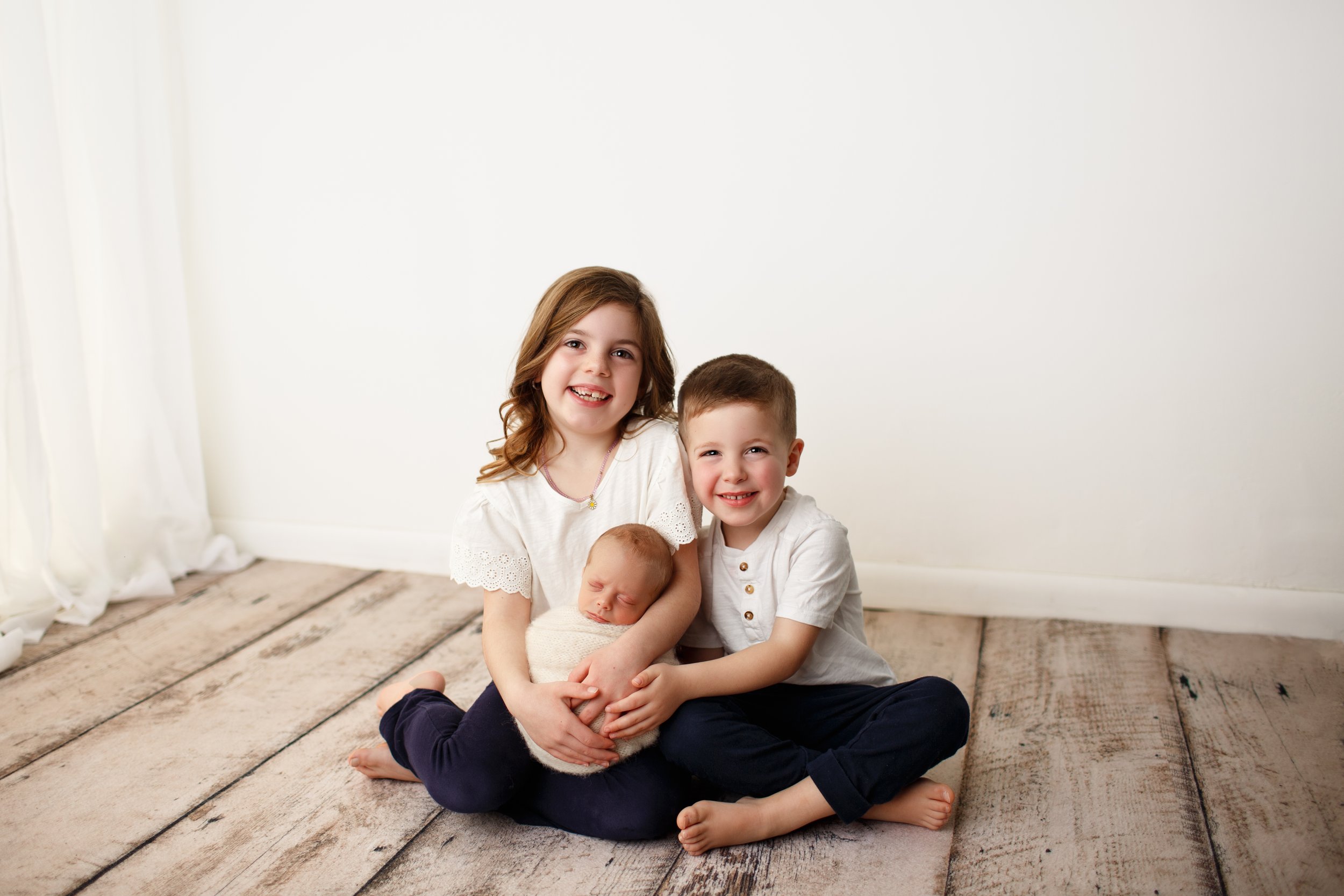  Siblings seated on wood floor holding their newborn sibling 