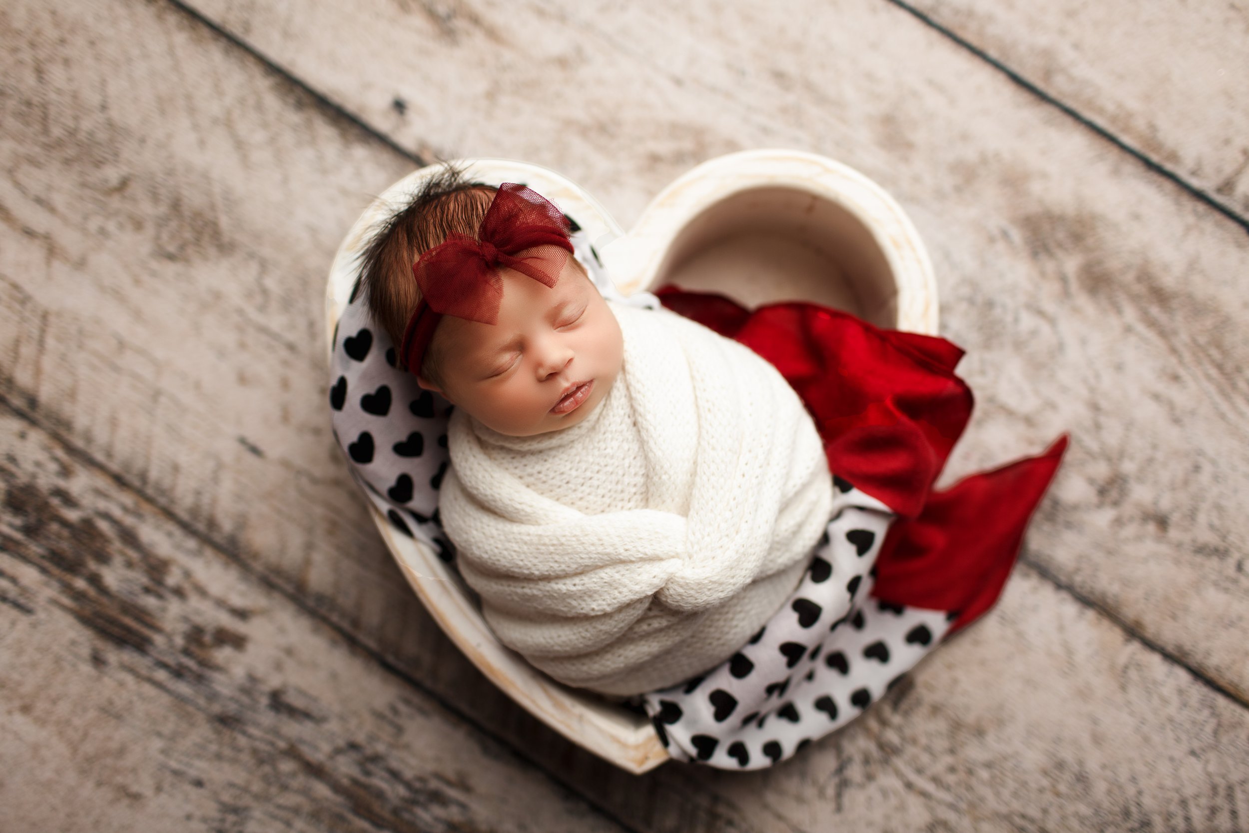  Baby girl in white swaddle lying in heart-shaped wooden bowl with a red blanket and a black and white polka-dot blanket next to her 