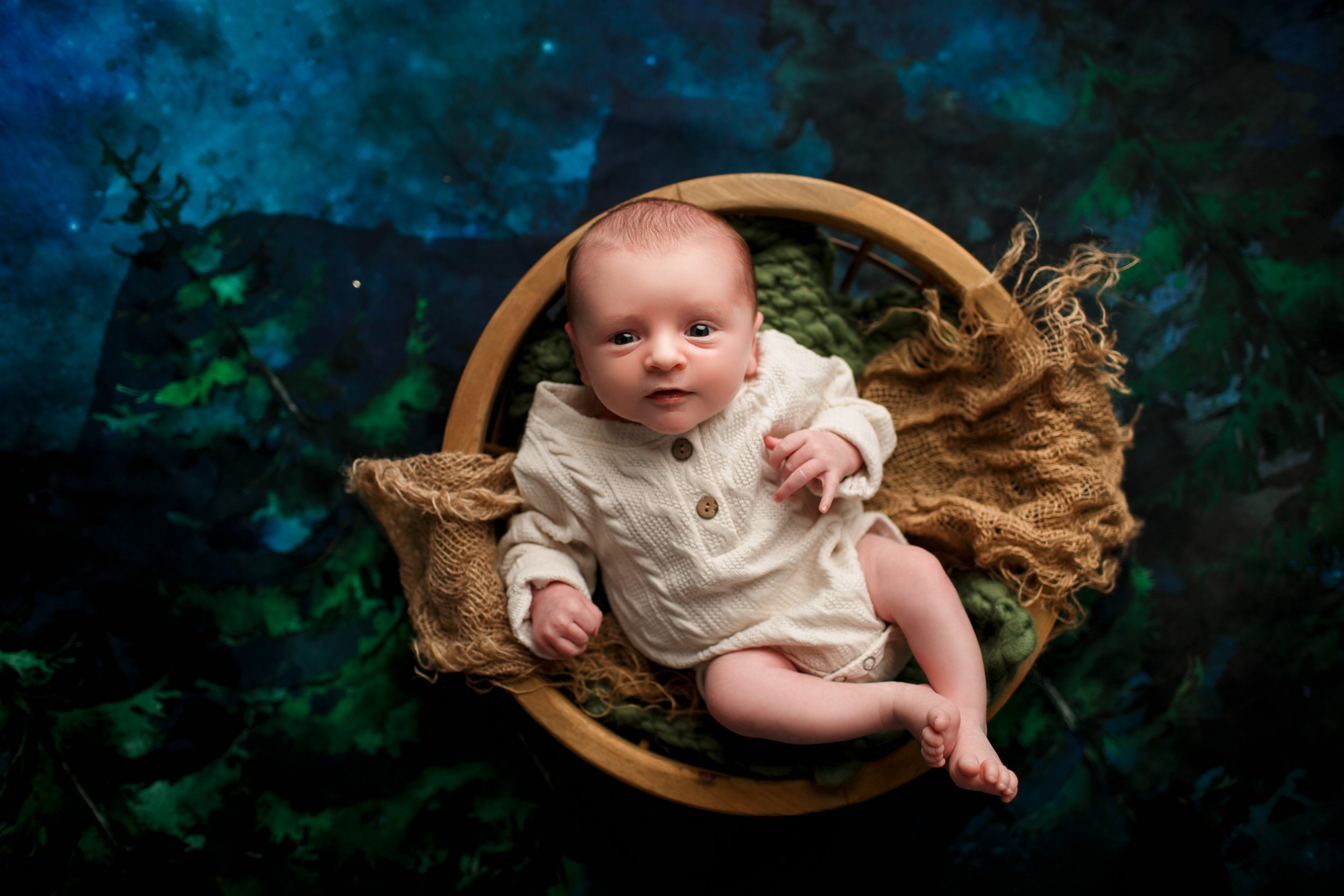  Wakeful baby boy in wooden bowl prop against a blue and green backdrop 