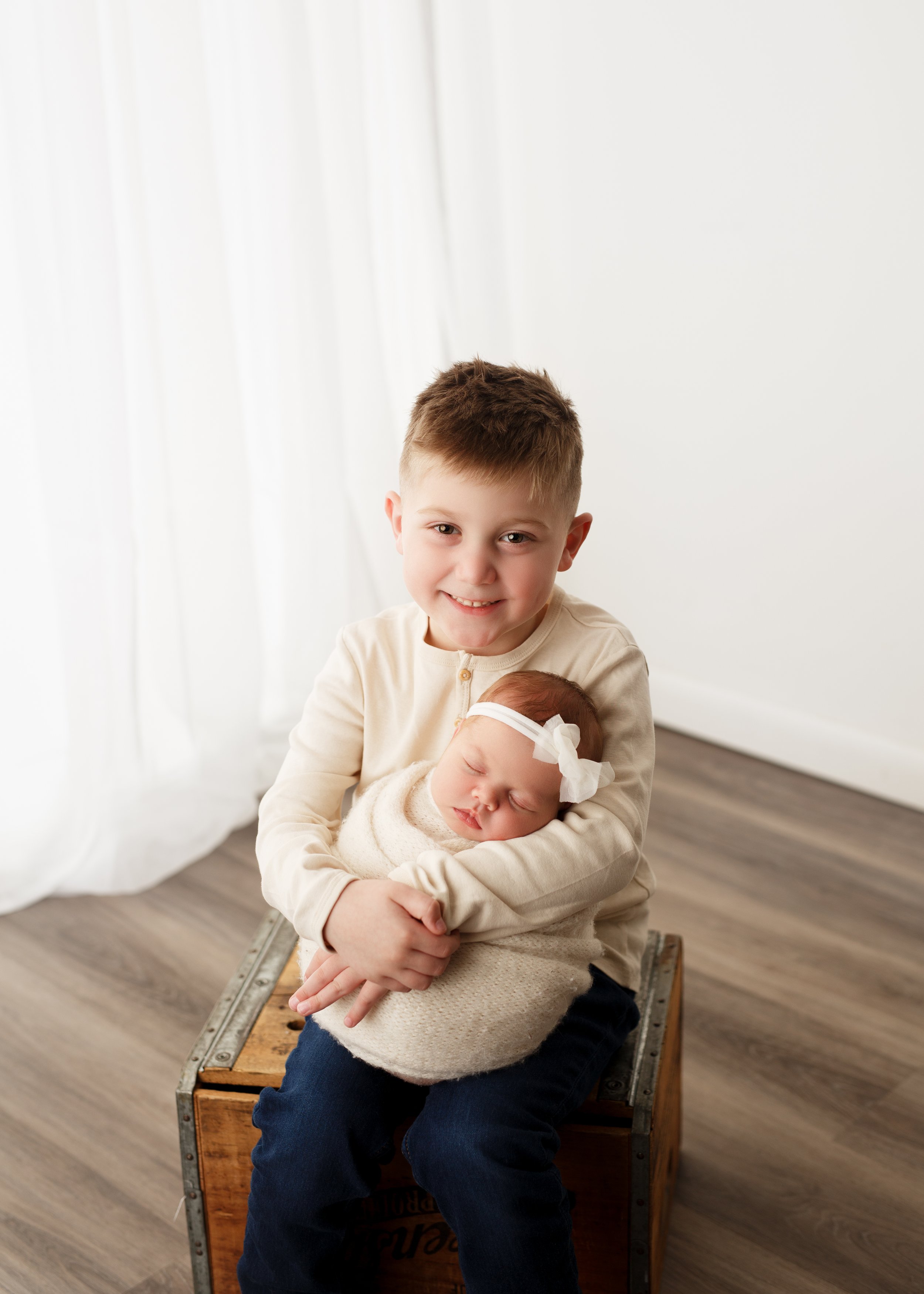  Little boy seated on a wooden crate holding his swaddled infant sister in his arms 