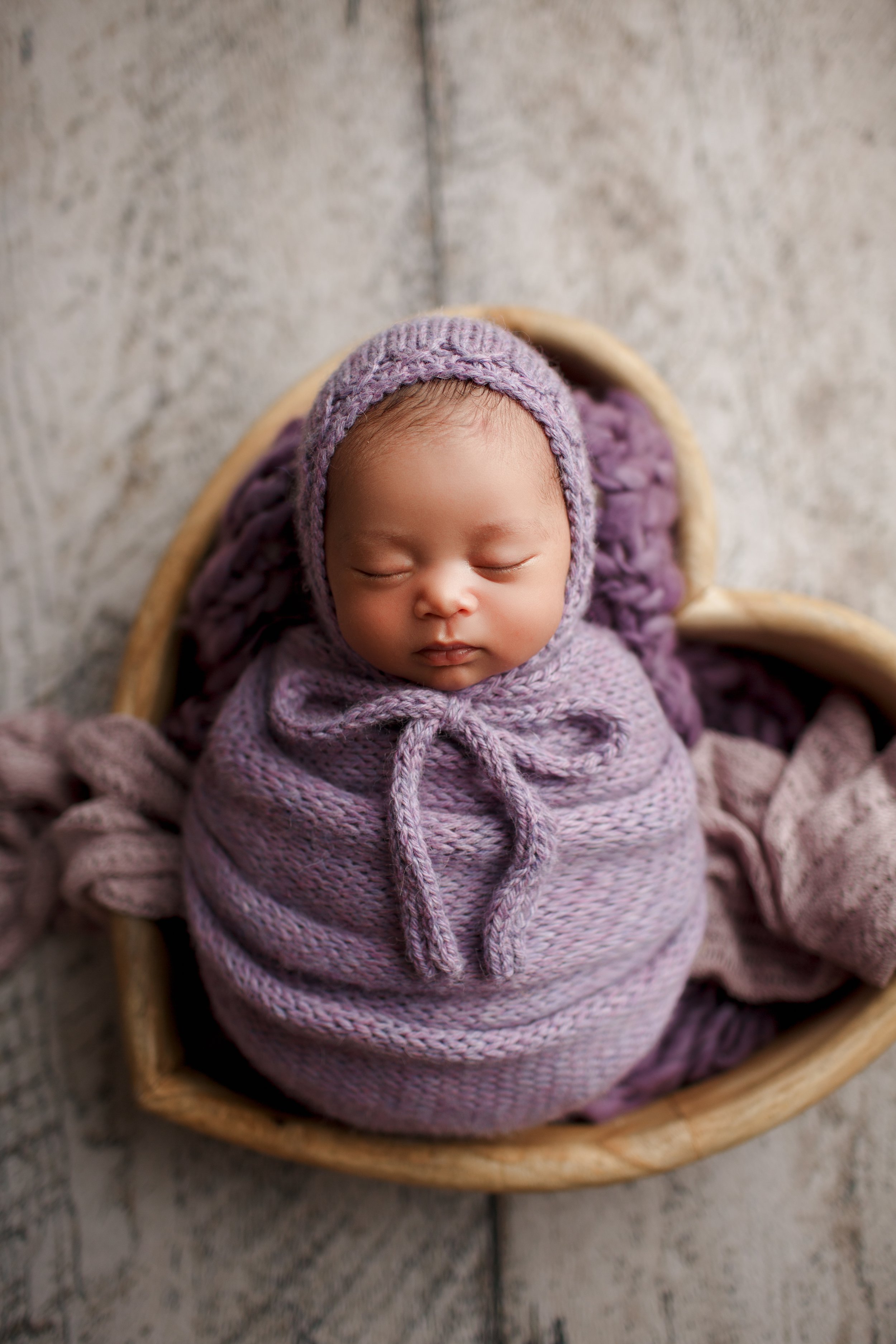  Baby in violet wrap and crochet beanie sleeps peacefully in wooden heart-shaped bowl 