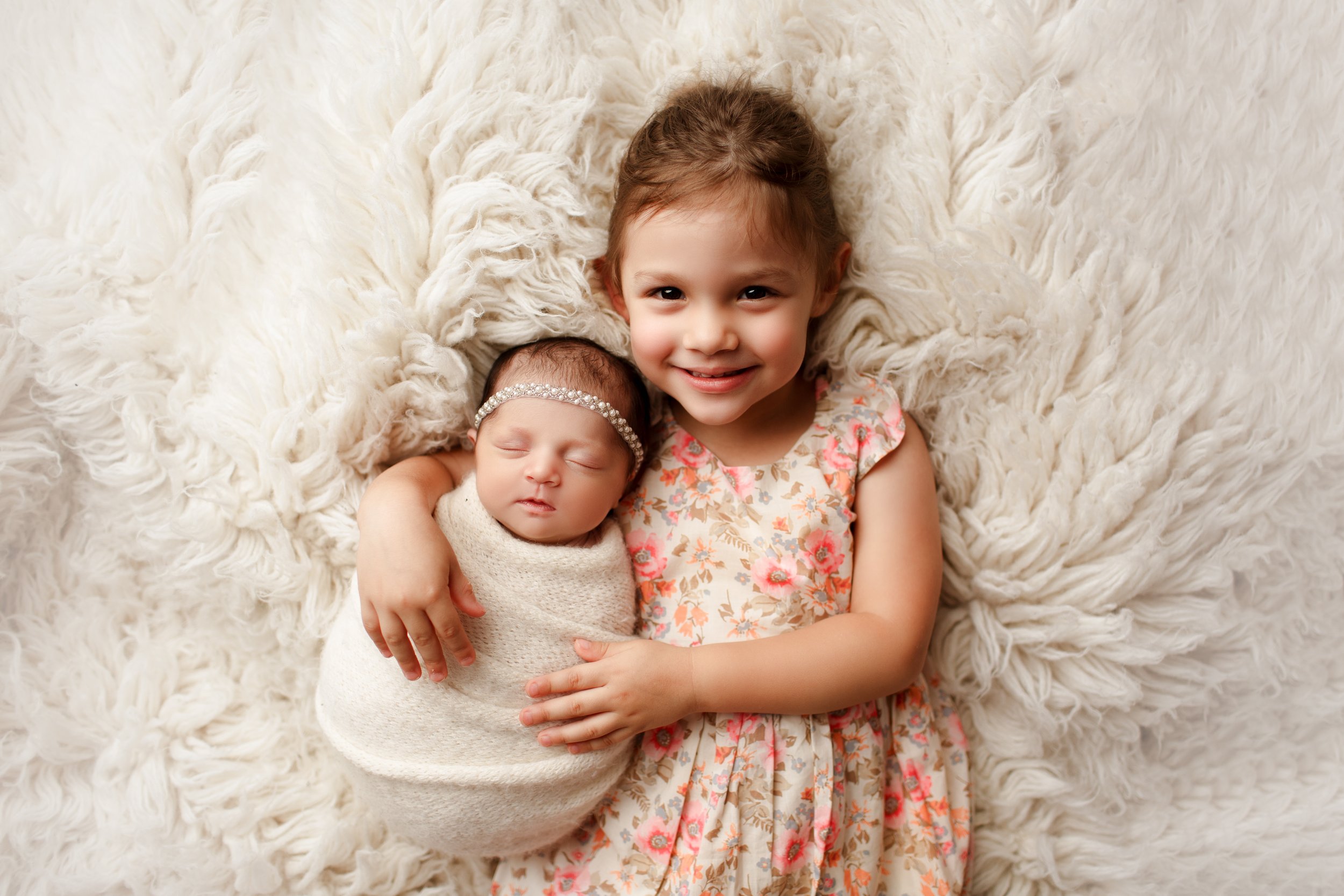  Little girl in floral print dress smiles happily as she and her new baby sister snuggle together on a faux fur rug  