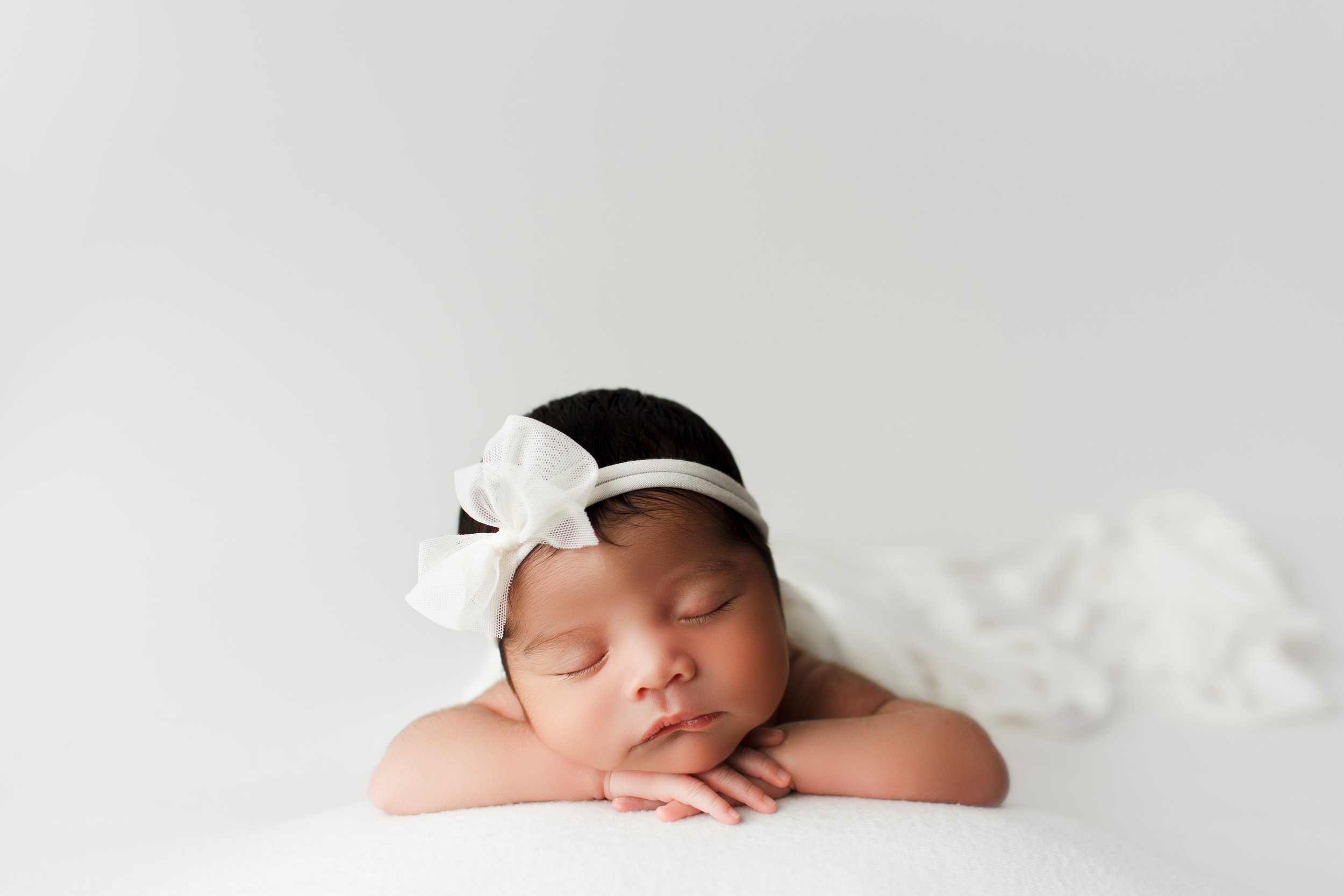  Dark-haired baby in white snoozes with her chin on her hands during her newborn portrait session 