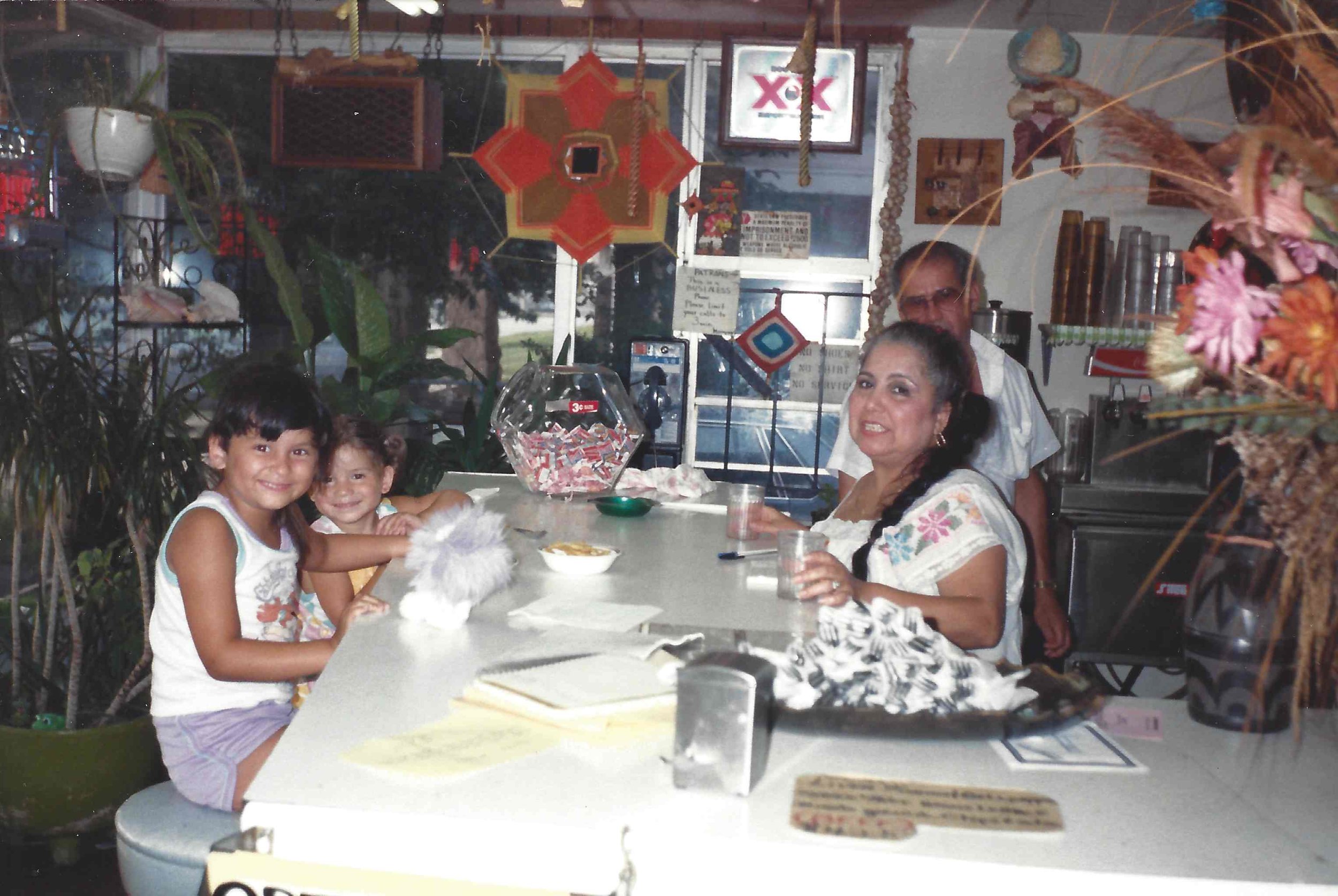 Gina (l) smiles while sitting at the booth with her sister Angelique (r) at their grandparents' restaurant Lupe and Bea's Texican restaurant in Memphis, Tennessee. Their grandmother Beatrice (l) and her friend Esteban Nodal Morales (r), a close friend who also helped at the restaurant, stand behind the booth. (Photo Courtesy: Angelique Sloan) 