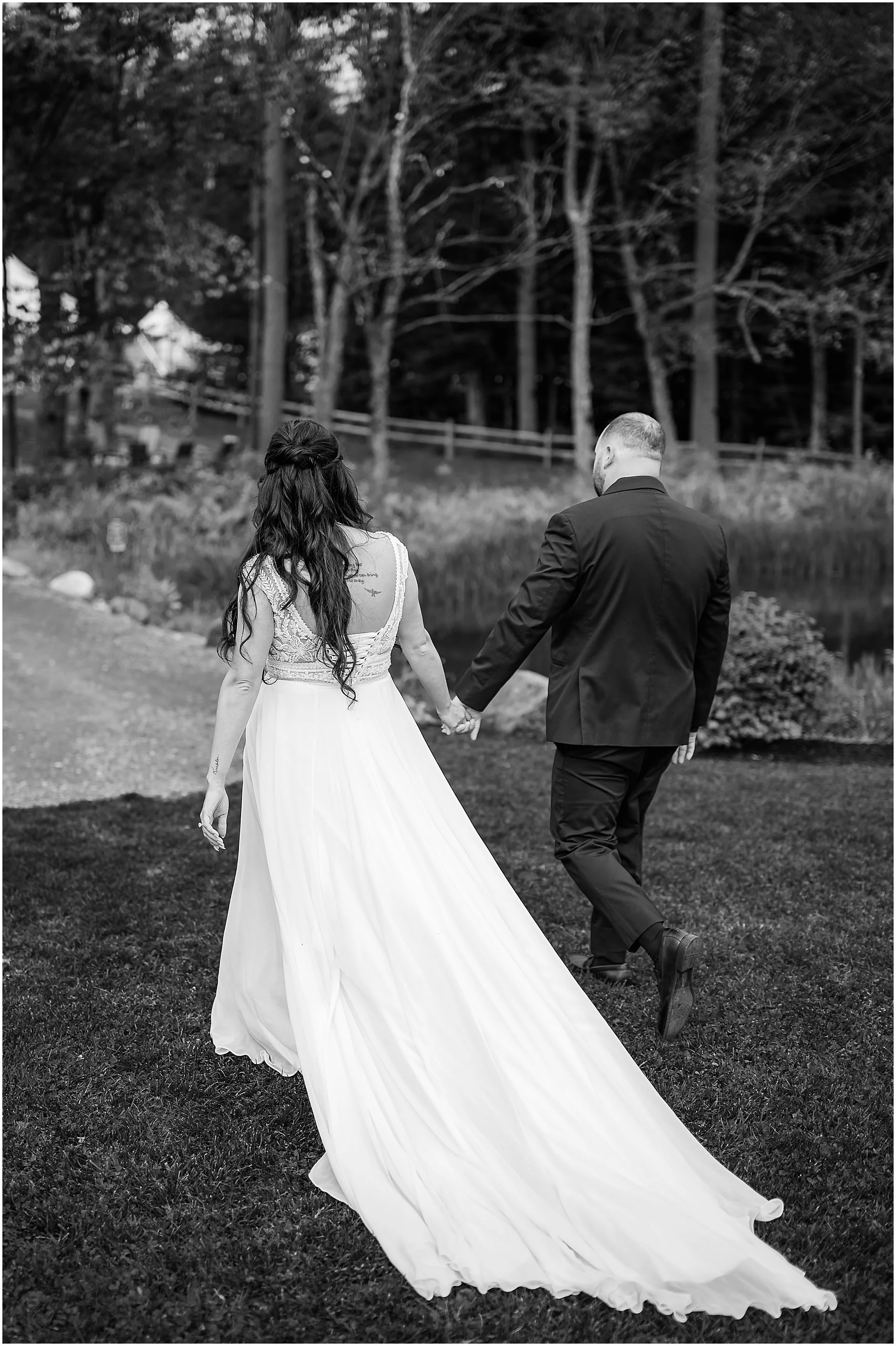 Black and white image of bride and groom hand in hand walking to the reception tent after their lakeside wedding ceremony