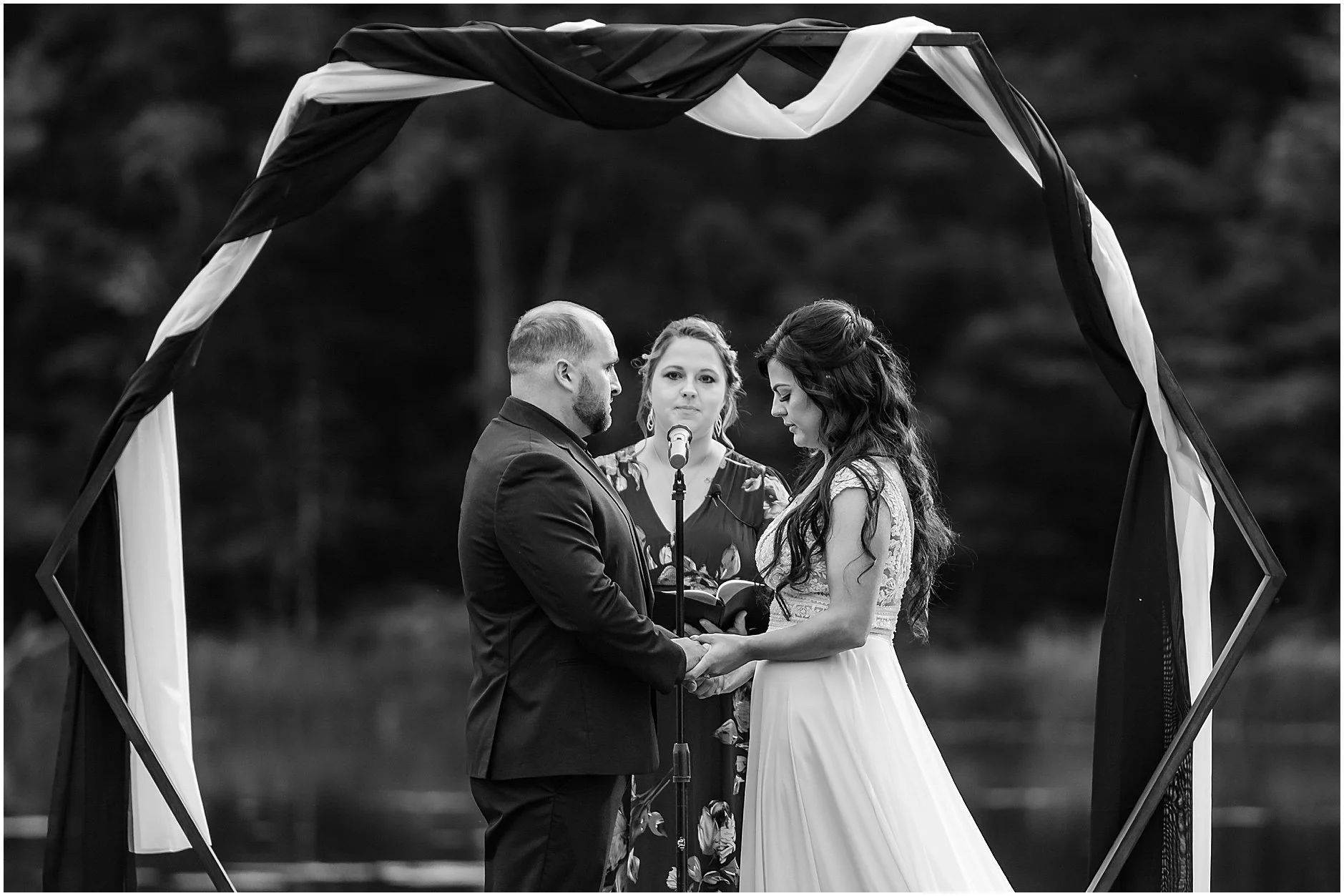 Intimate black and white image of bride and groom holding hands during lakeside wedding ceremony