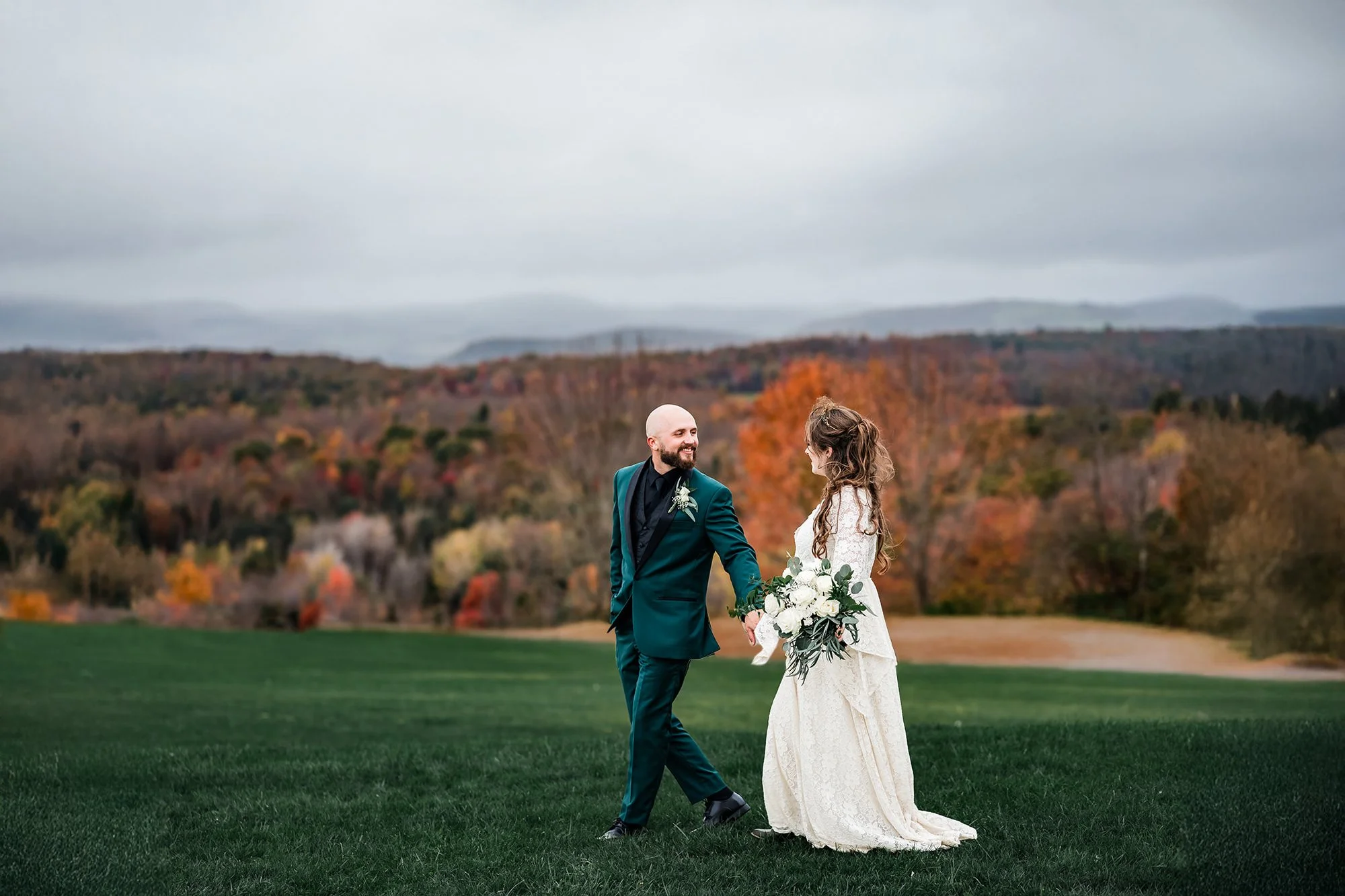 Bride and groom holding hands and smiling outdoors on their wedding day, with autumn trees and mountains in the background.