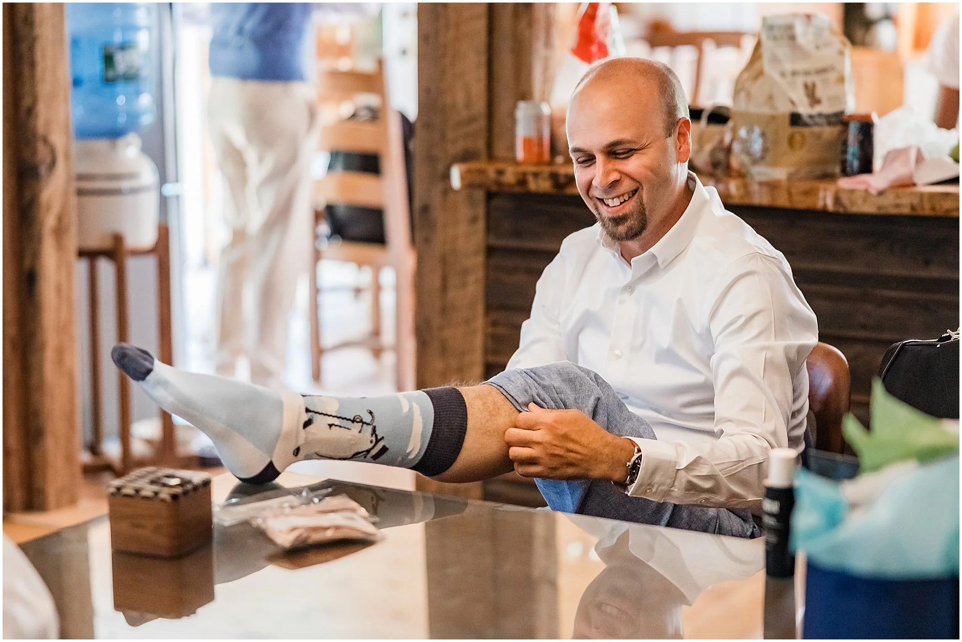 Groom putting on his socks as he smiles getting ready for his wedding at The Inn at West Settlement