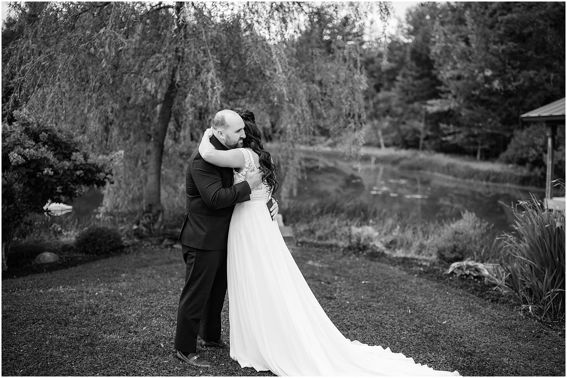 Black and white image of bride and groom sharing a hug during their first look under a willow tree at The Rosemary at Spano Lake in Cooperstown