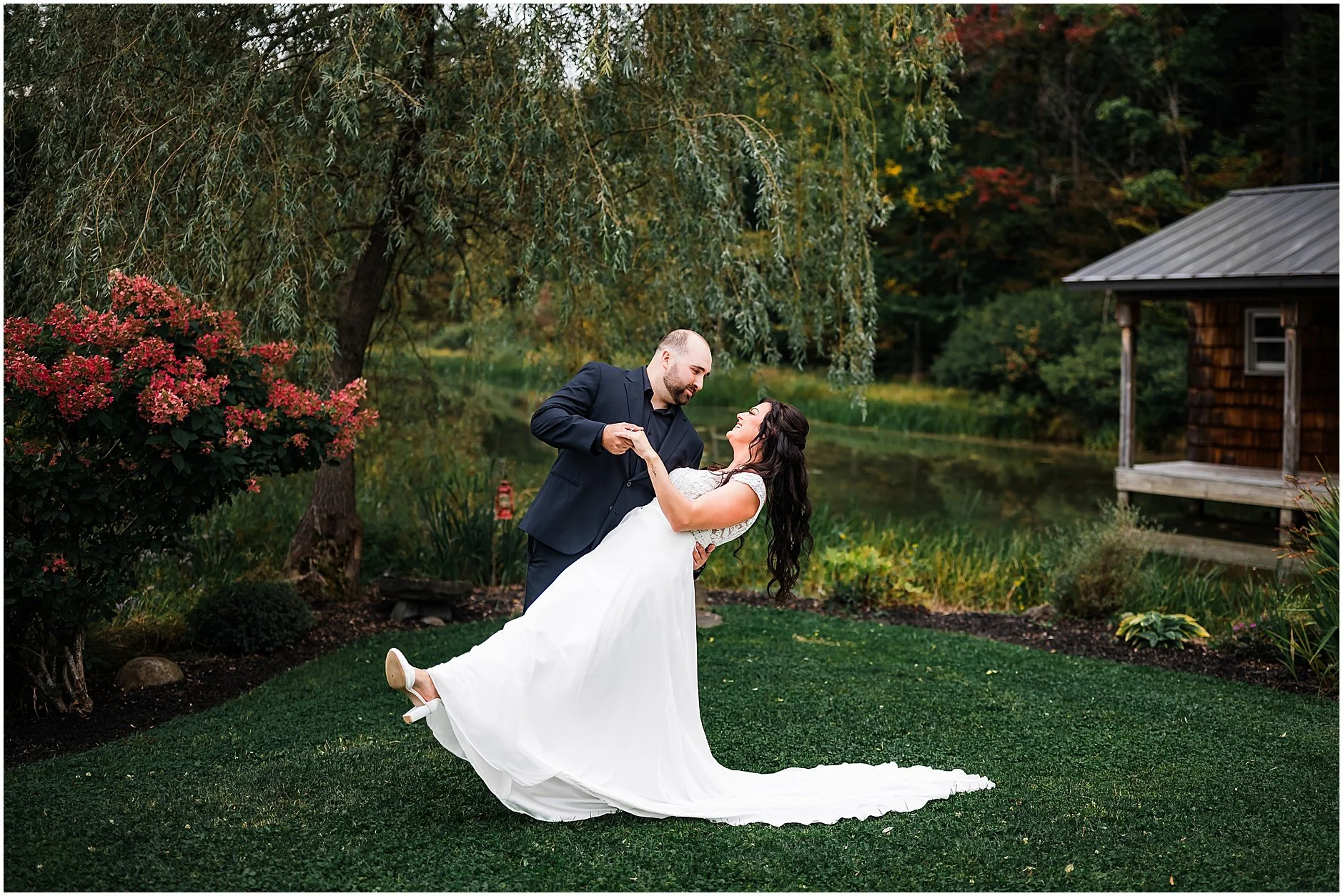 Groom dipping his bride under a willow tree by the water at The Rosemary at Spano Lake in Cooperstown New York
