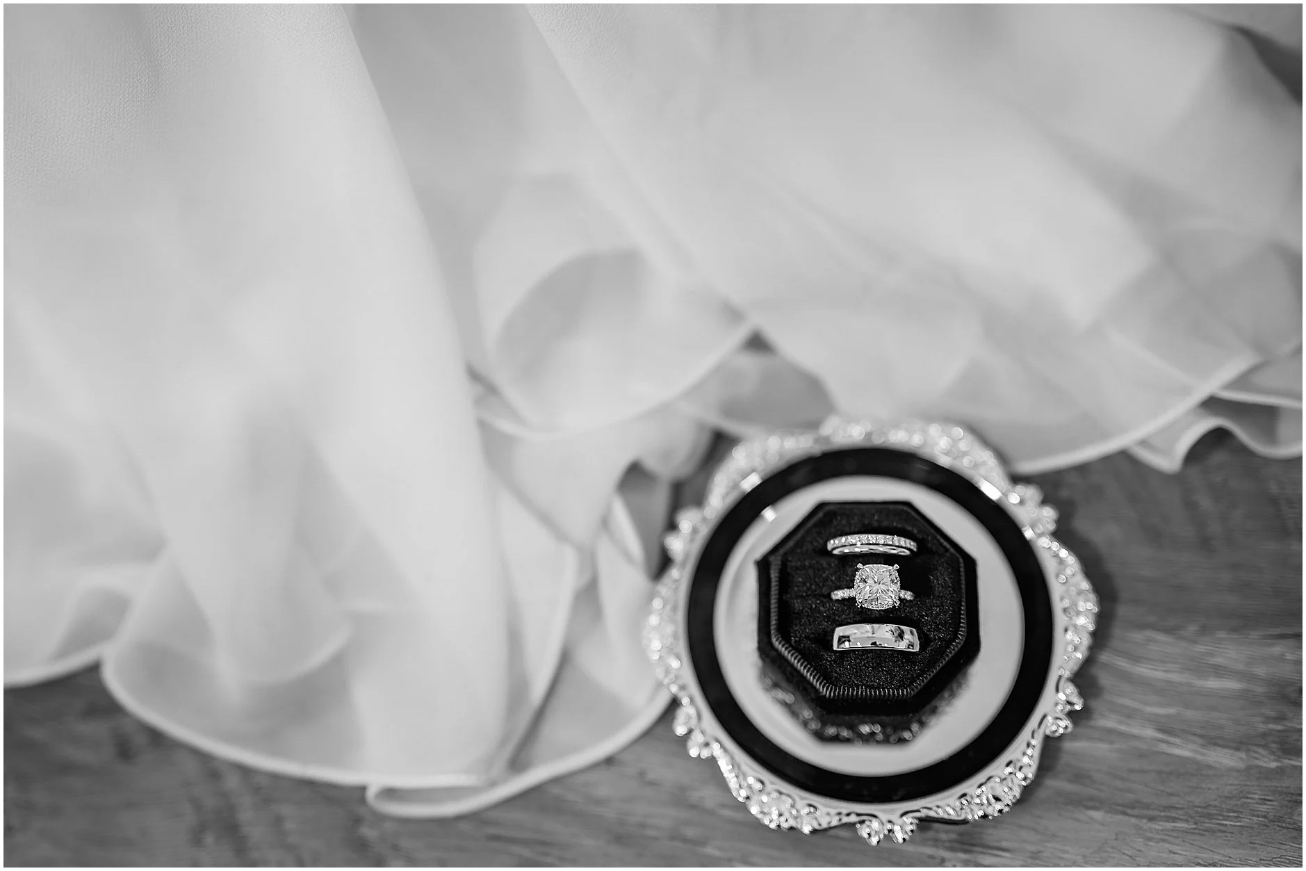 Black and white image of rings in ring box against bride's white wedding dress