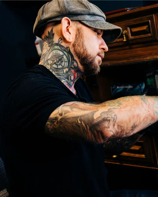 Side profile of a man wearing a gray cap, tattoos on his neck and arms, and a black shirt, sitting indoors in front of wooden cabinets.