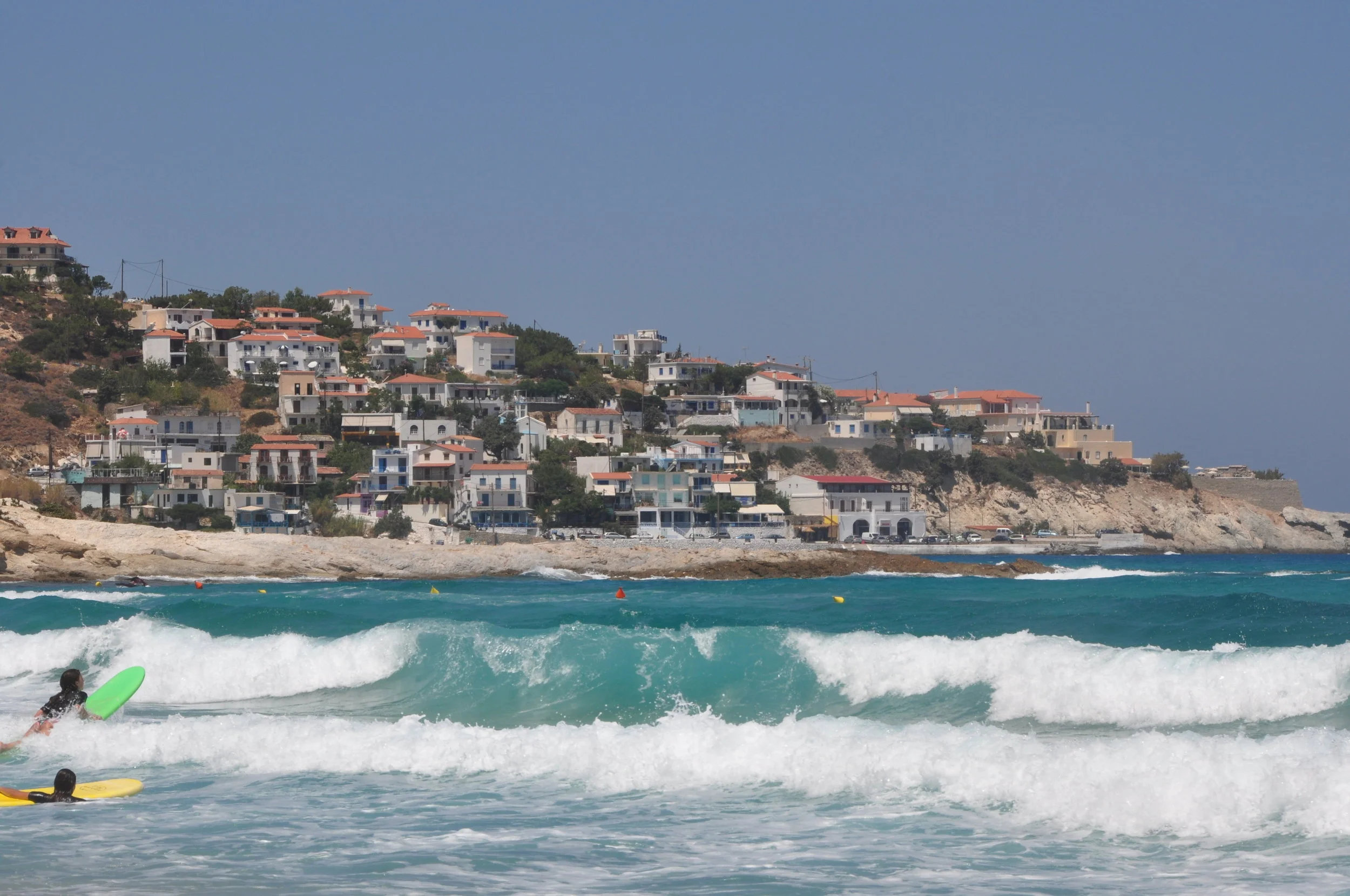 Surfers paddling out at Mesakti Beach, Ikaria, with a view of Armenistis village