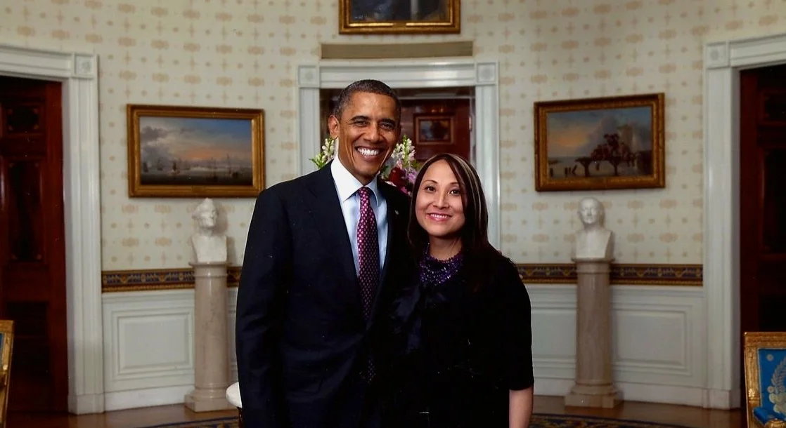 Former President Barack Obama and activist Cecilia Chung stand side-by-side, smiling, in the Oval Office of the White House. President Obama is on the left, wearing a dark suit, white shirt, and a red patterned tie. Cecilia Chung is on the right, wearing a dark top with a sparkling purple neckline. Behind them is the distinctive curved wall of the Oval Office, featuring framed paintings and white marble busts on pedestals.
