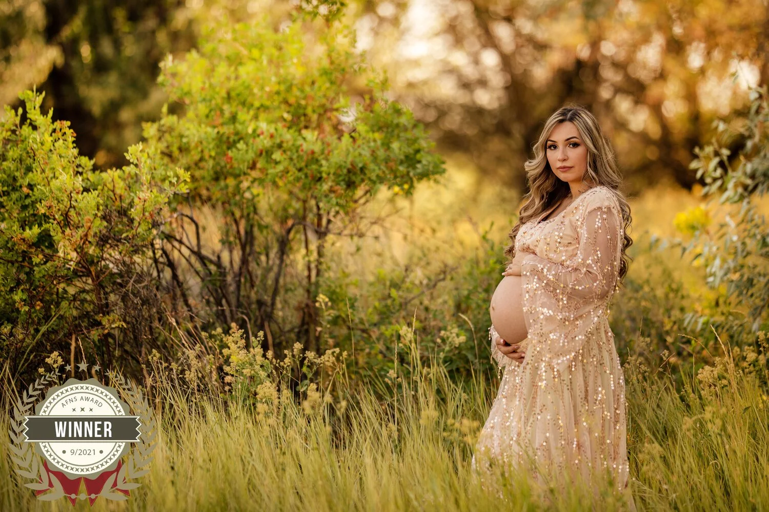 Pregnant woman in a beige sequined dress standing in a grassy field with autumn trees in the background, holding her belly, with a "Winner" badge in the lower left corner.