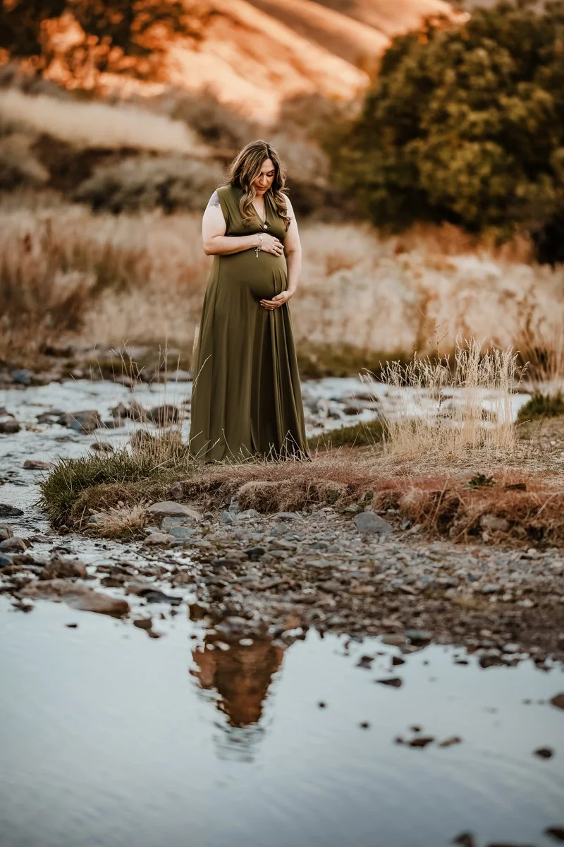 Pregnant woman in a green dress stands near a river, gently cradling her belly with both hands amidst a natural outdoor setting with trees and hills in the background.