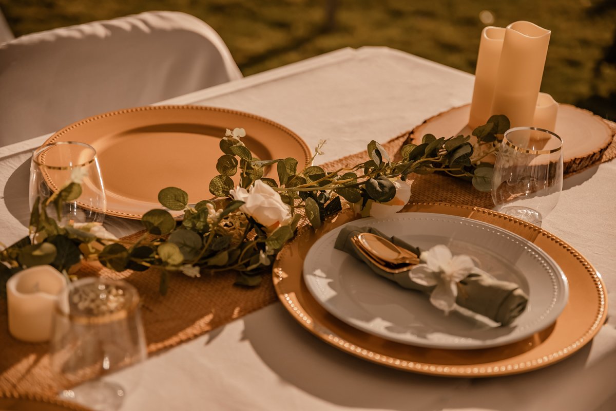 Elegant table setting with gold and white dinnerware, a floral garland, candles, and glassware, set on a white tablecloth for a special occasion.