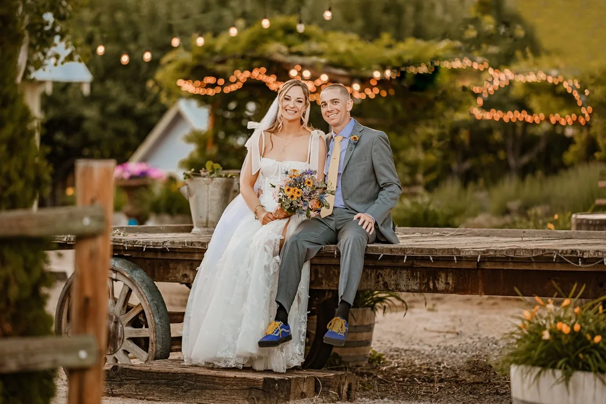 A bride and groom sitting on a rustic wooden cart outdoors during their wedding, with string lights and trees in the background.