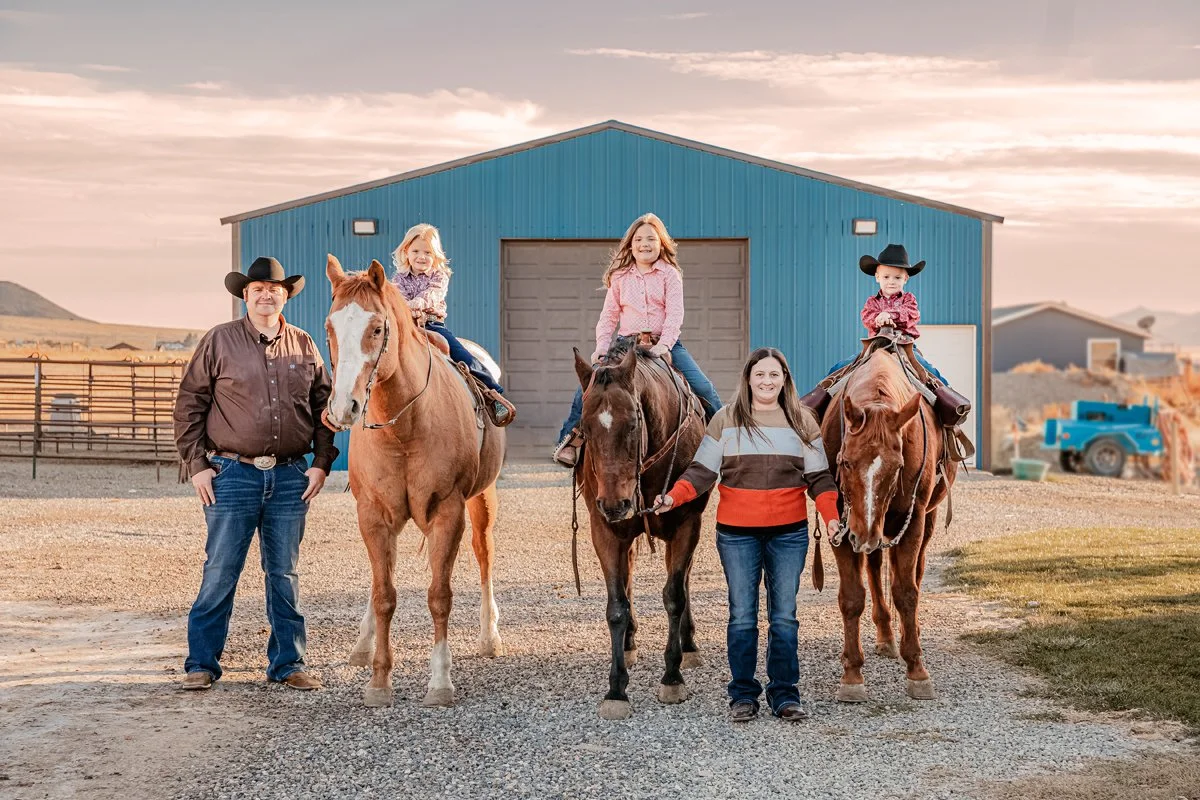 A family of five with two children riding horses on a farm in front of a blue barn, at sunset.