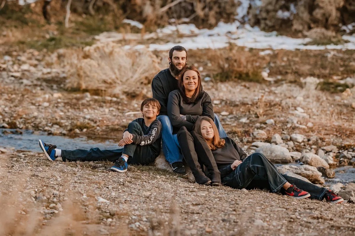 A family of four sitting and lying on a rocky riverbank outdoors. The father and mother sit close together with two children, one girl leaning against her mother and a boy lying on the ground nearby, all enjoying a relaxed moment in nature.