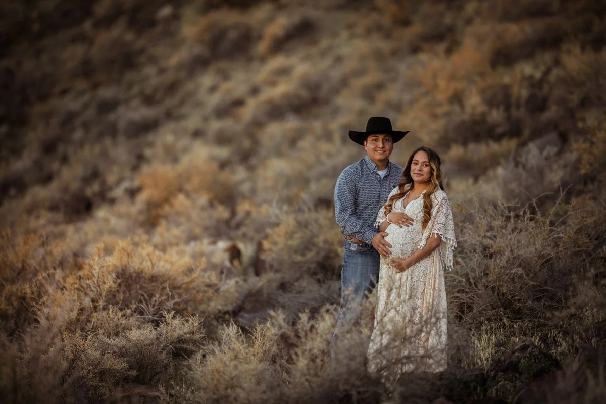 A young couple, with the woman being pregnant, standing in a desert landscape with dry shrubs and brown rocks, smiling at the camera. The man is wearing a black cowboy hat, and the woman is wearing a white lace dress.