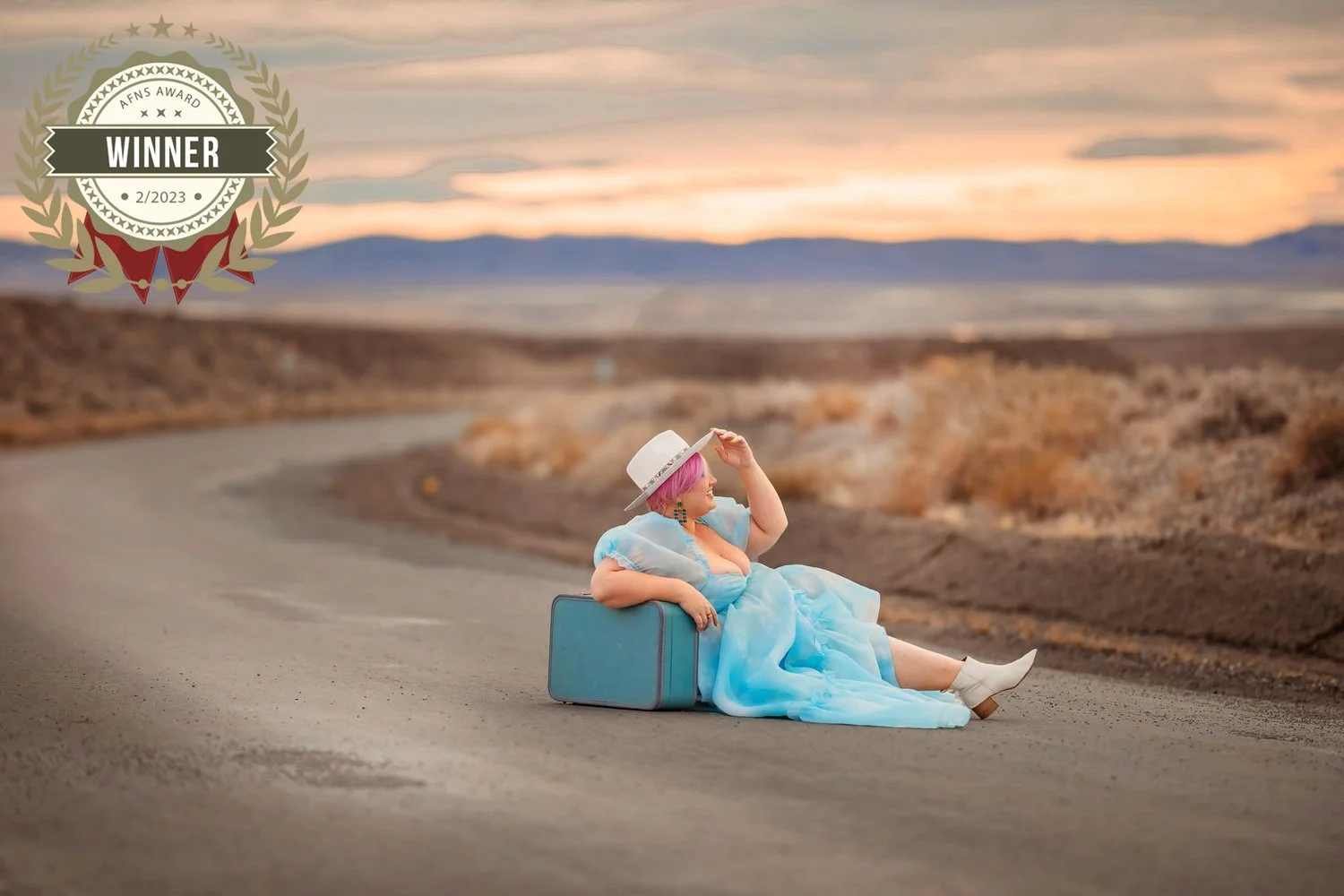 Woman with pink hair wearing a blue dress, white boots, and a white hat sitting on a suitcase on a deserted road at sunset, smiling and looking into the distance.