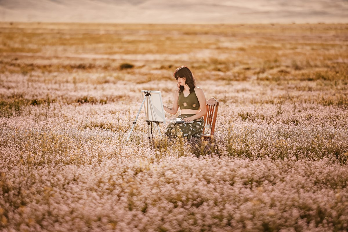 A woman sitting on a wooden chair in a field of pink and white flowers, painting on a canvas set up on an easel during sunset.