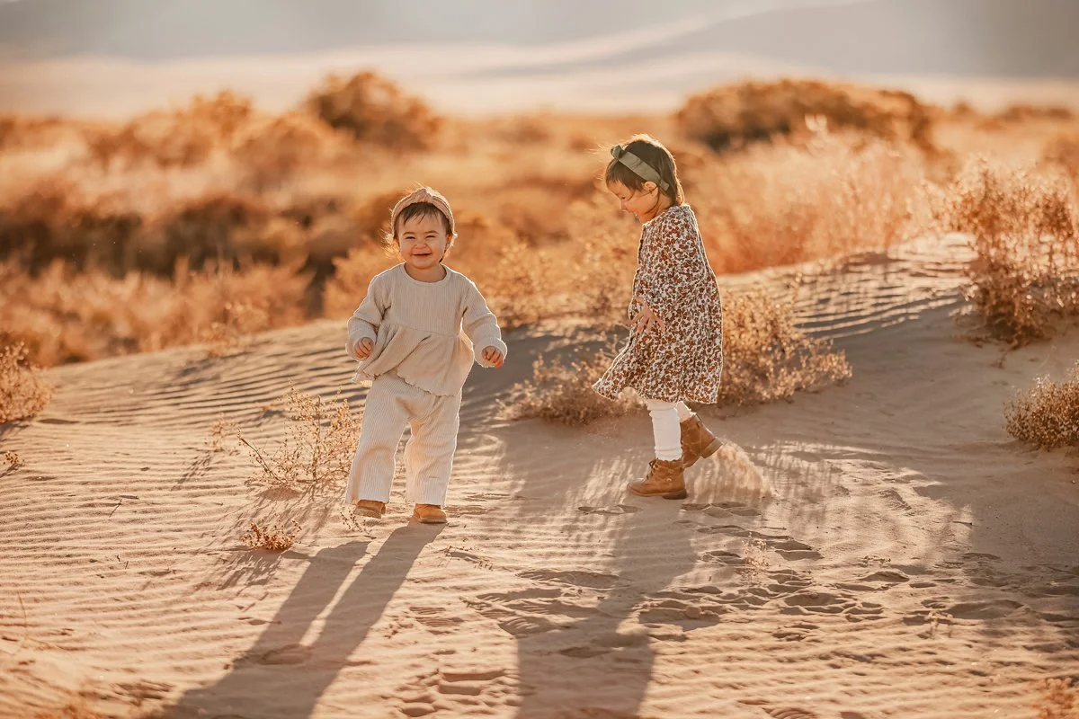 Two young girls playing and smiling in a sandy desert landscape, surrounded by dry bushes, with the sun shining warmly.