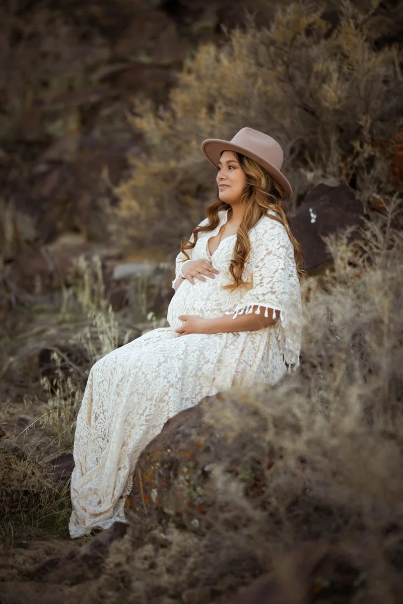 Pregnant woman sitting on rocks in outdoor natural setting, wearing a white lace dress and a wide-brimmed beige hat, holding her belly with a serene expression.
