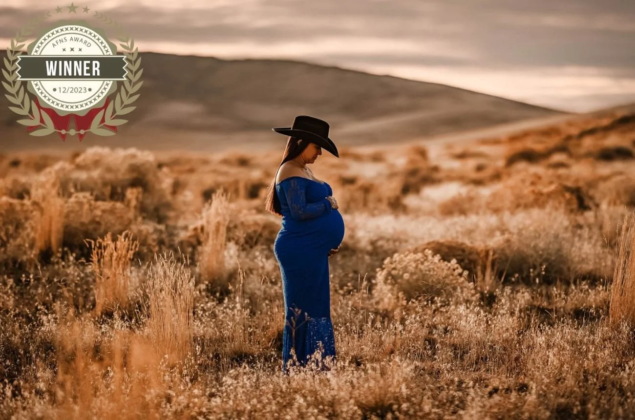 A pregnant woman in a blue dress and black hat standing in a field of dry grass during sunset.