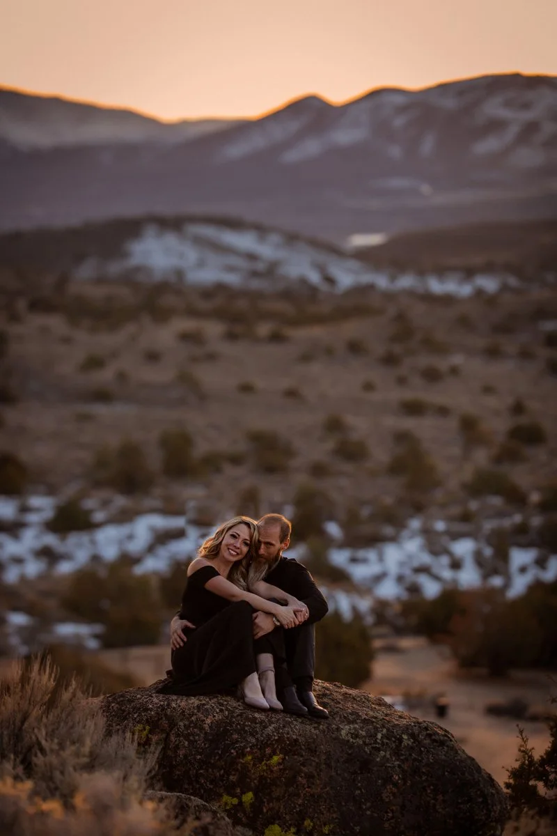 A couple sitting together on a large rock in a mountainous desert landscape during sunset, embracing and smiling.