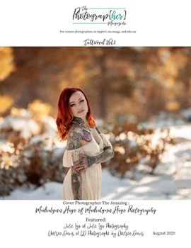Portrait of a woman with red hair and tattoos standing outdoors in autumn foliage, arms crossed, smiling.