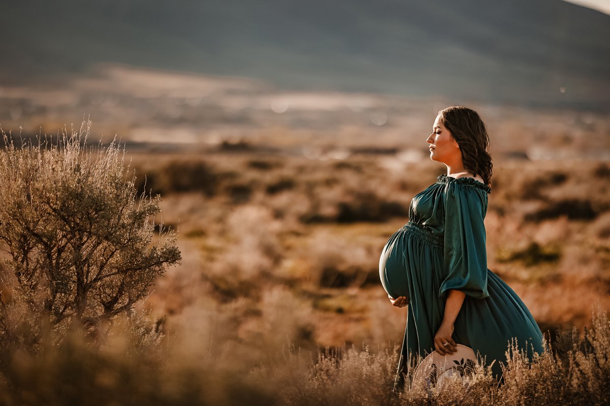 Pregnant woman in a teal dress standing in a desert landscape with mountains in the background.