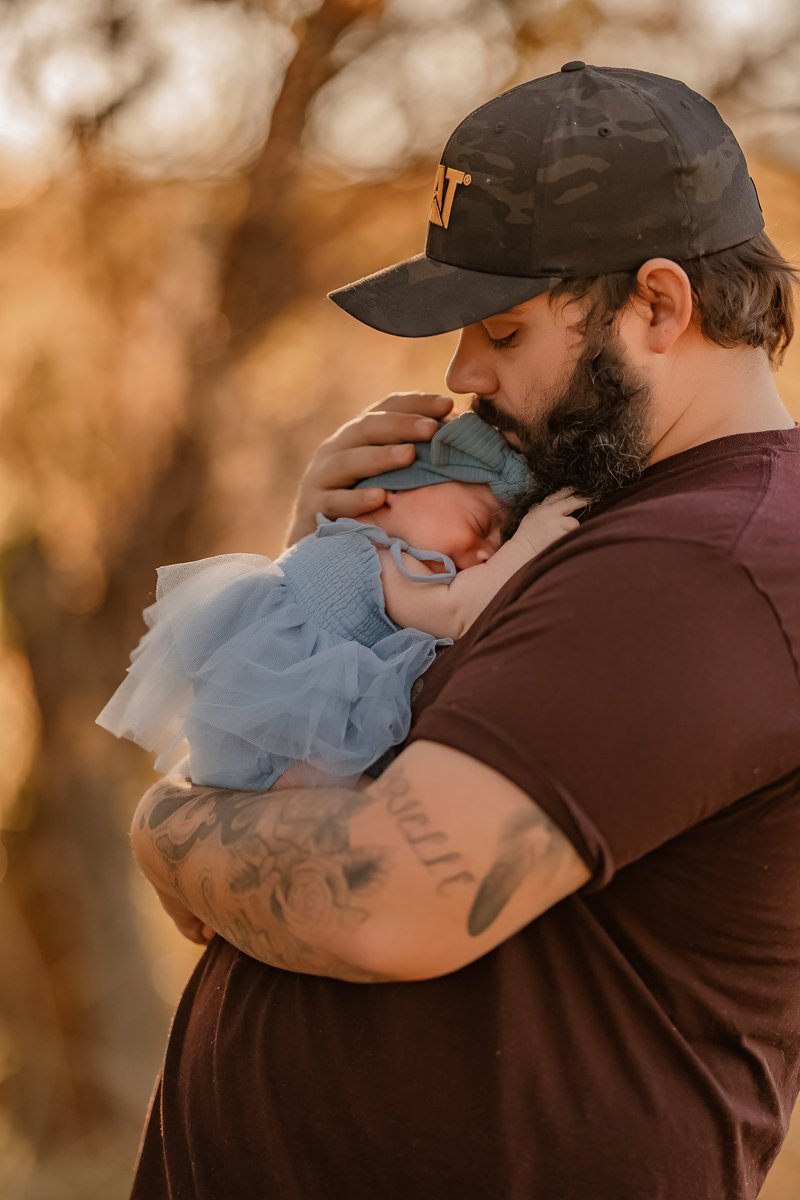 A man holding a sleeping baby close to his face outdoors during autumn. The man has tattoos on his arm and a beard, wearing a cap and a dark t-shirt.