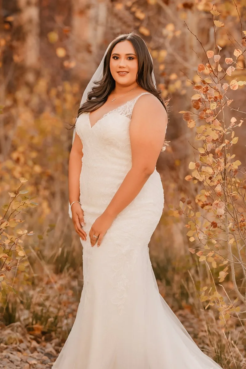 A woman in a white wedding dress standing outdoors among autumn foliage, smiling at the camera.