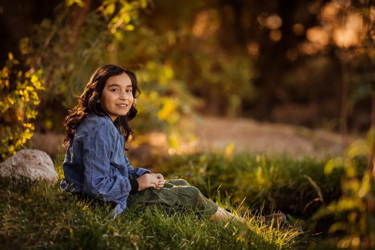A young girl sitting on grass near a small stream during sunset, smiling at the camera, surrounded by trees and nature.