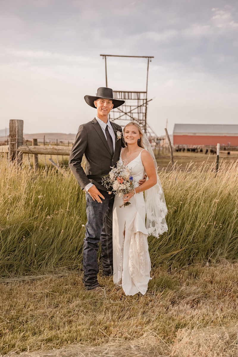 A couple dressed in wedding attire standing in a grassy field with a rustic barn and a wooden scaffolding in the background during sunset.